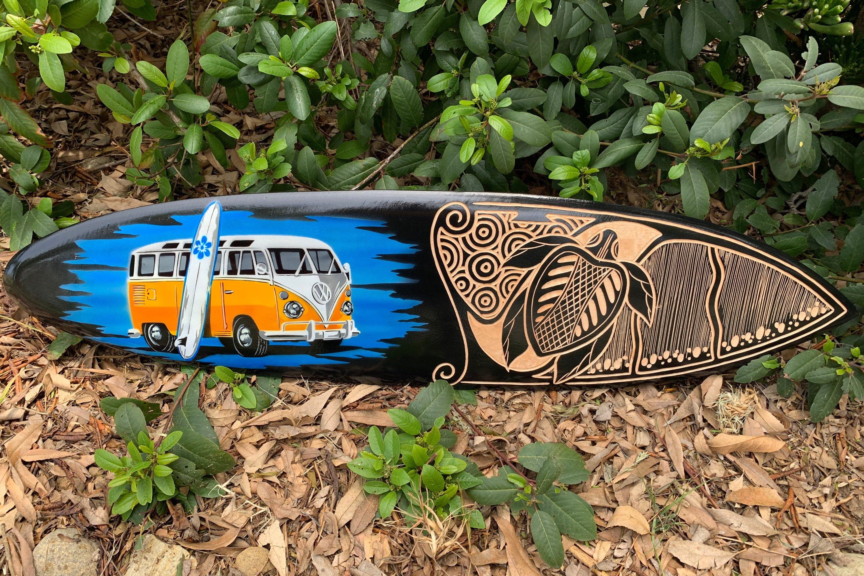 Decorated surfboard with an orange VW bus and a tiki motif on a black background