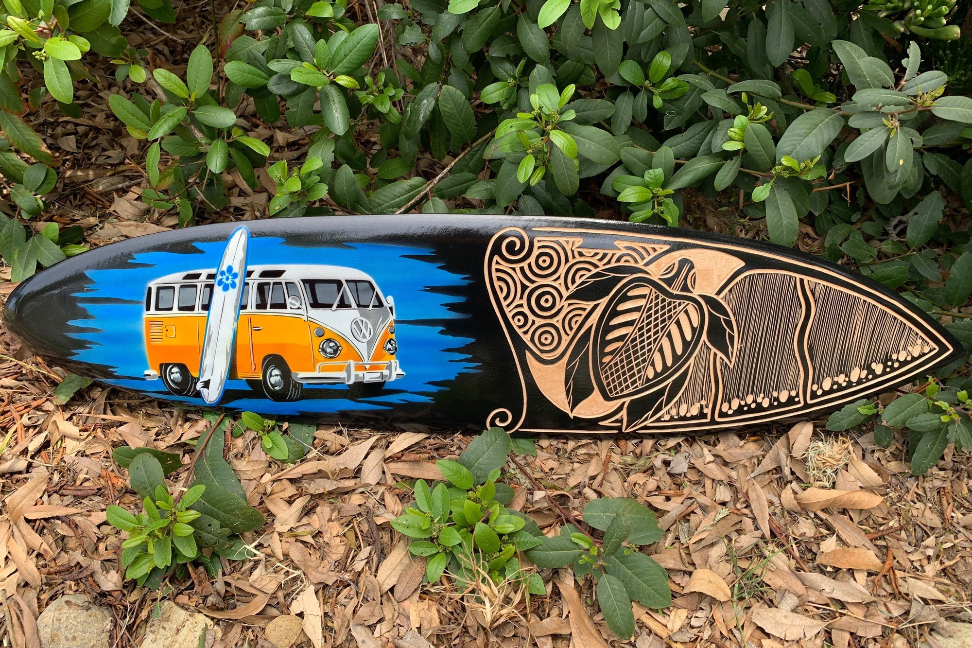 Decorated surfboard with an orange VW bus and a tiki motif on a black background
