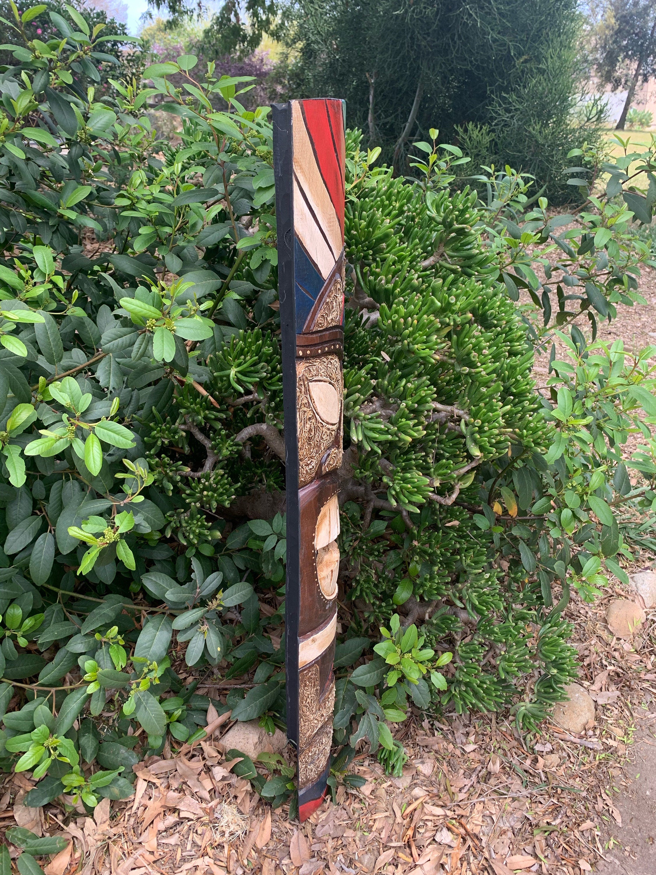 Tall decorative painted wooden totem-style sculpture leaning against a green shrub in a garden.