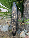 Black surfboard with beige floral and geometric pattern leaning against a tree outdoors