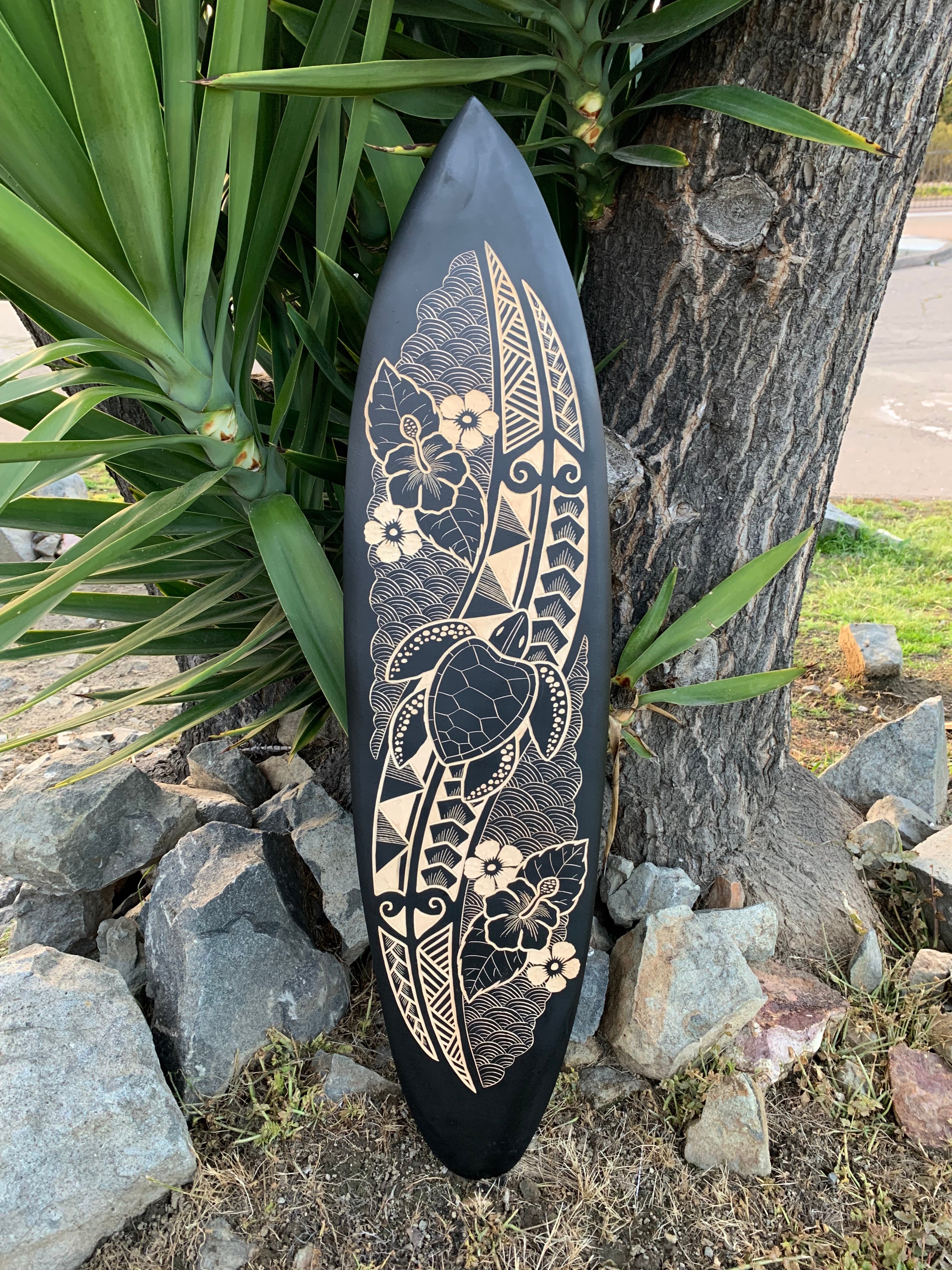Black surfboard with a beige floral and turtle design leaning against a tree