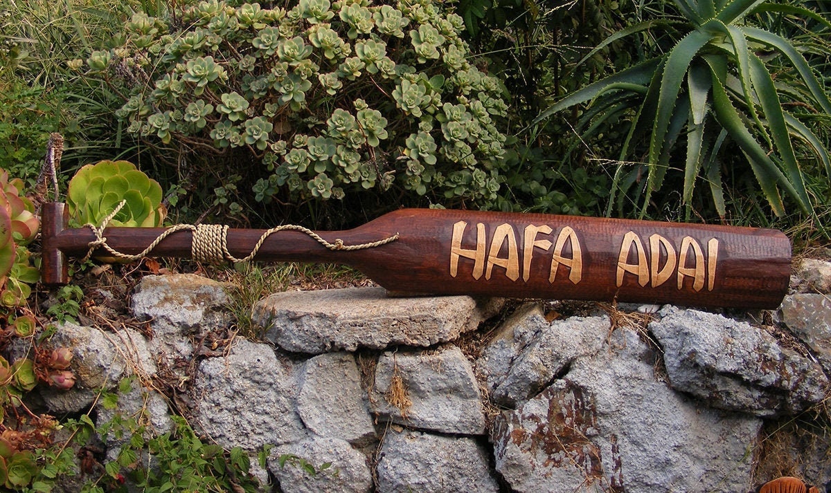 Carved wooden bat reading 'HAFA ADAI' placed on a stone wall with succulents