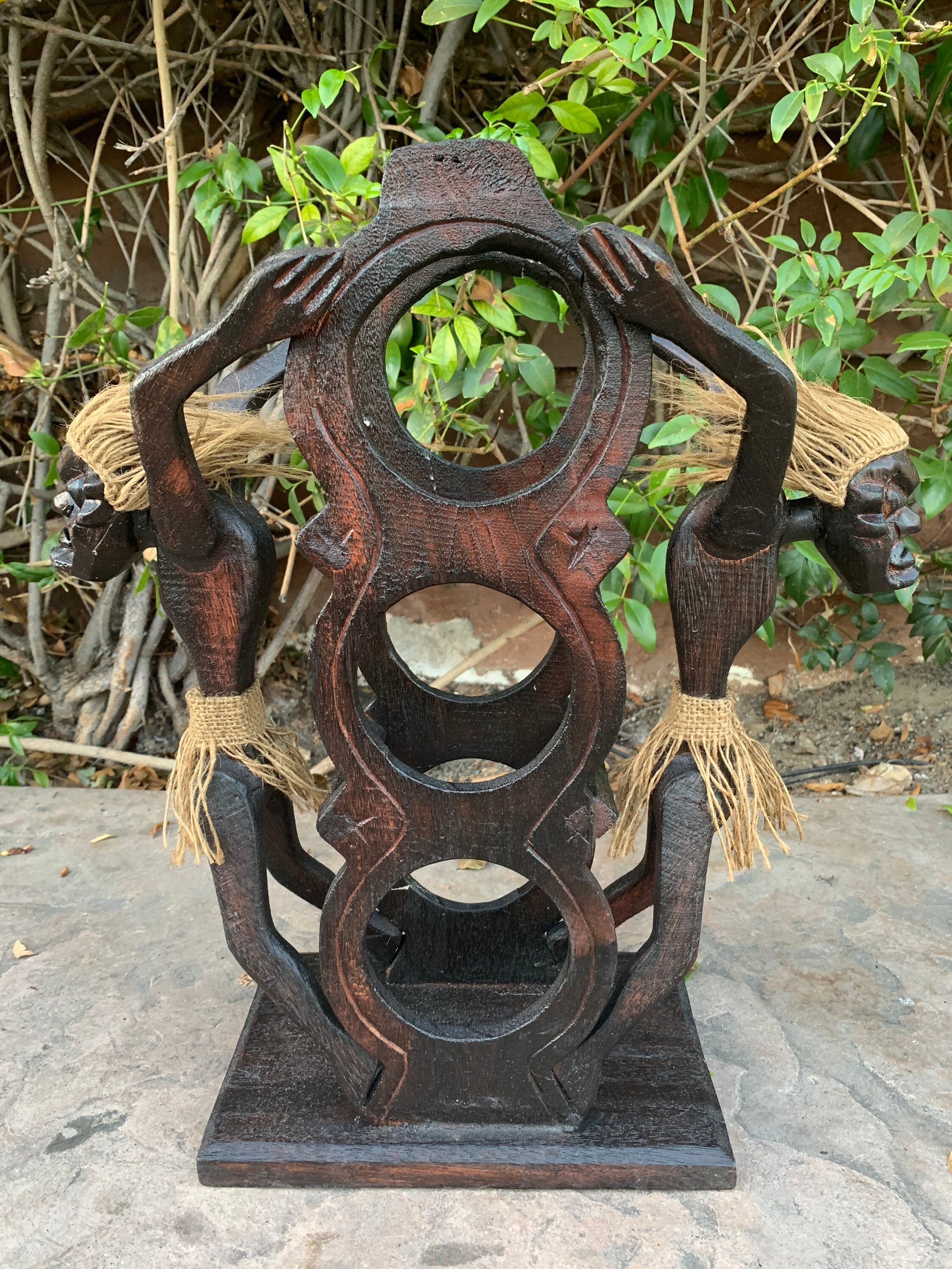 Dark hand-carved wooden sculpture with circular cutouts and jute tassels in a garden setting.