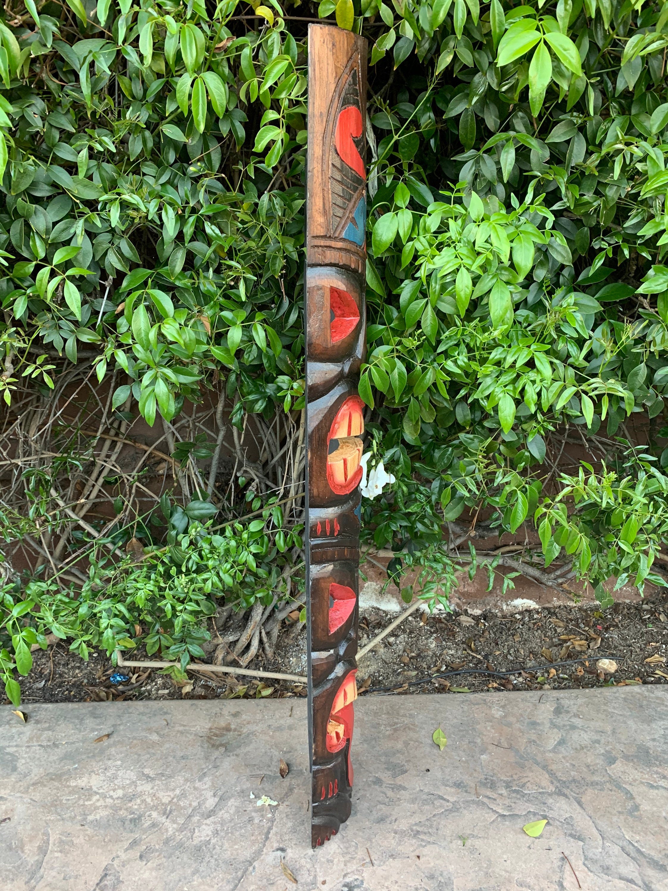 Hand-carved wooden totem pole with red and orange inlays standing beside green bushes in a garden.