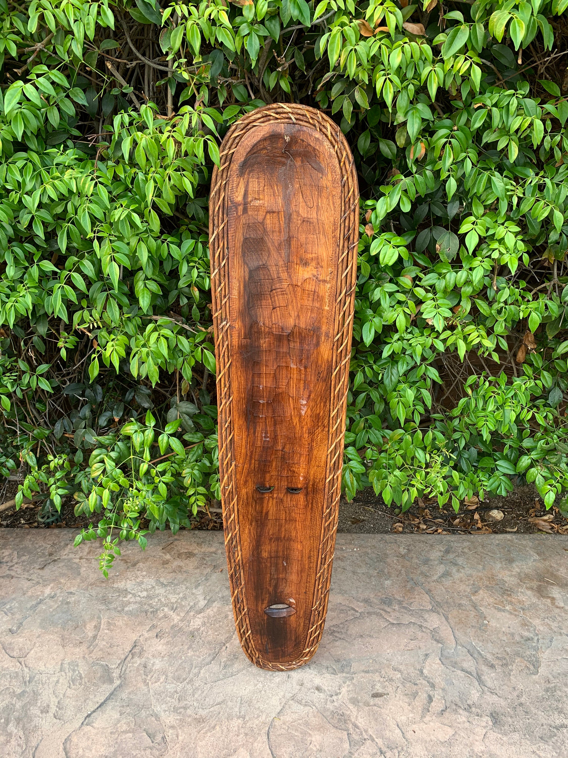 Long wooden carved board with rope edging leaning against a green hedge