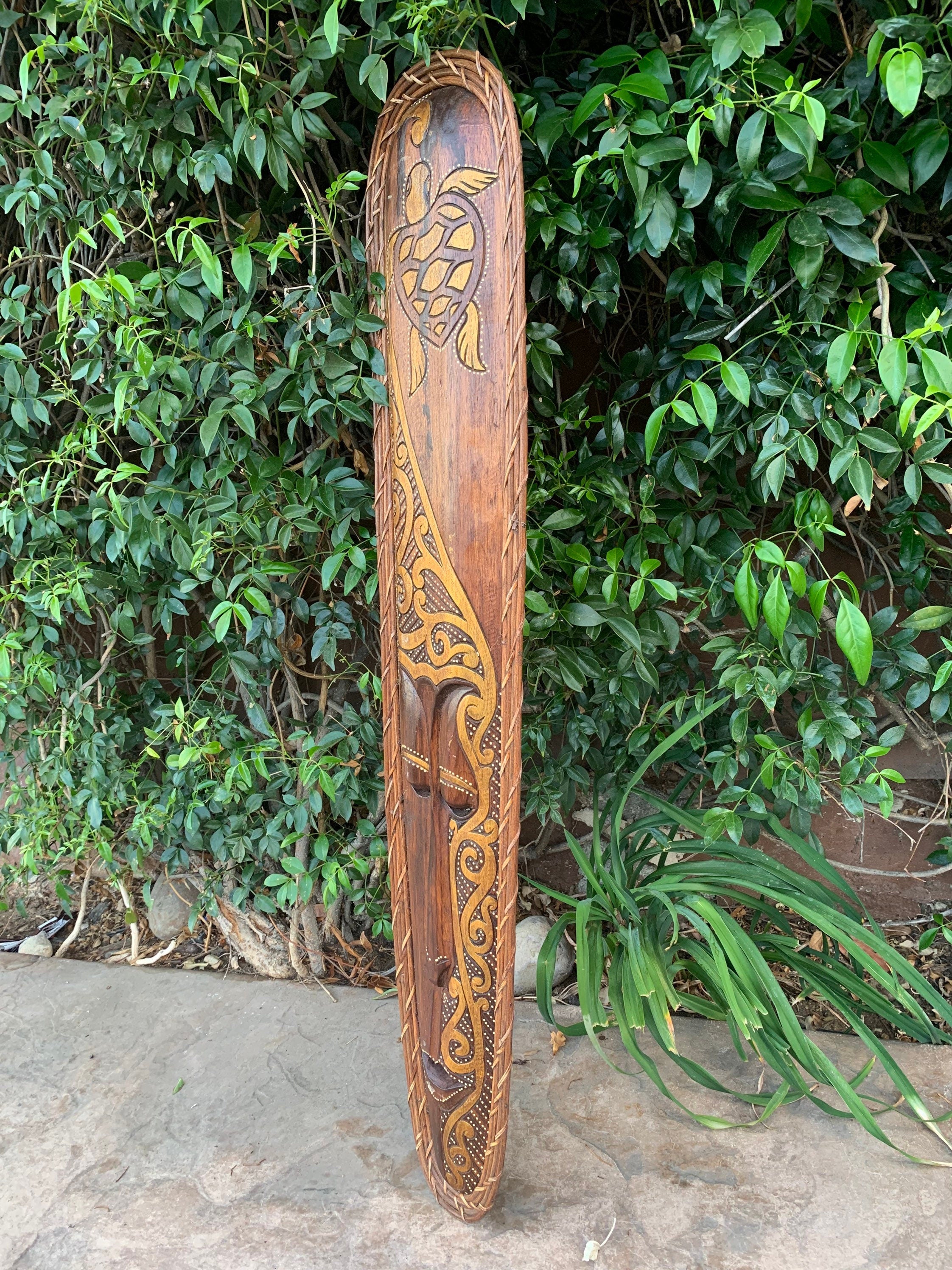Hand-carved wooden paddle with a turtle motif and decorative scrollwork leaning against greenery