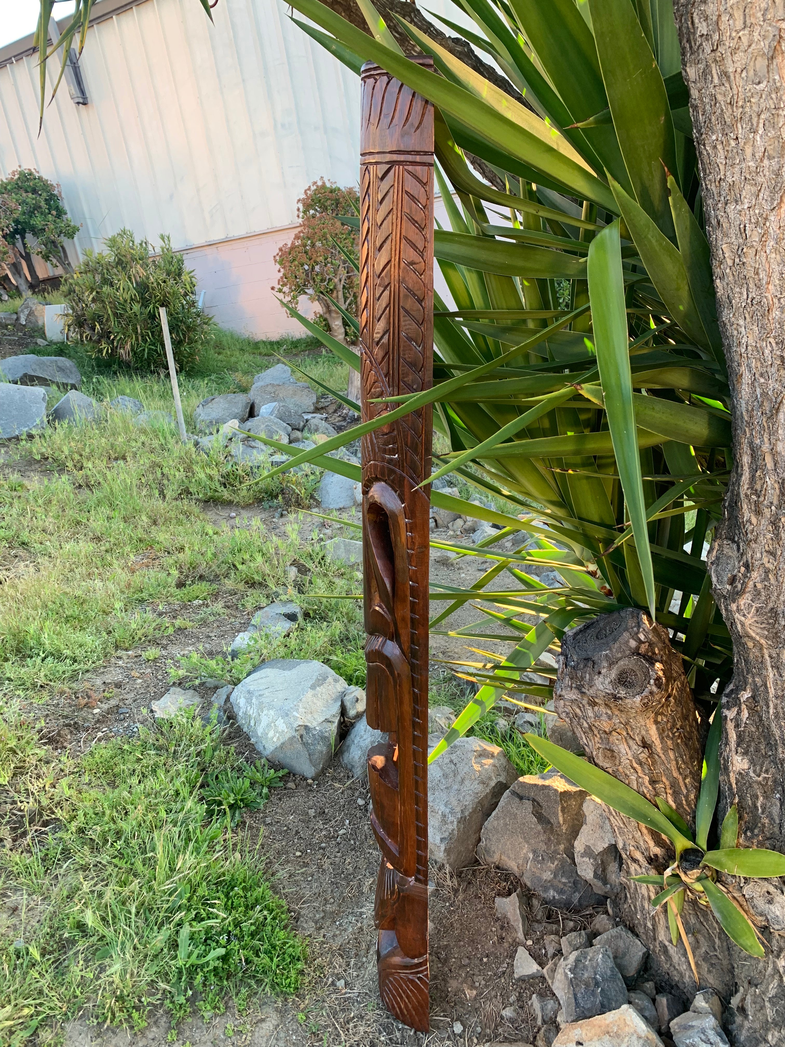 Hand-carved wooden post with chevron patterns leaning beside a tree in a garden.