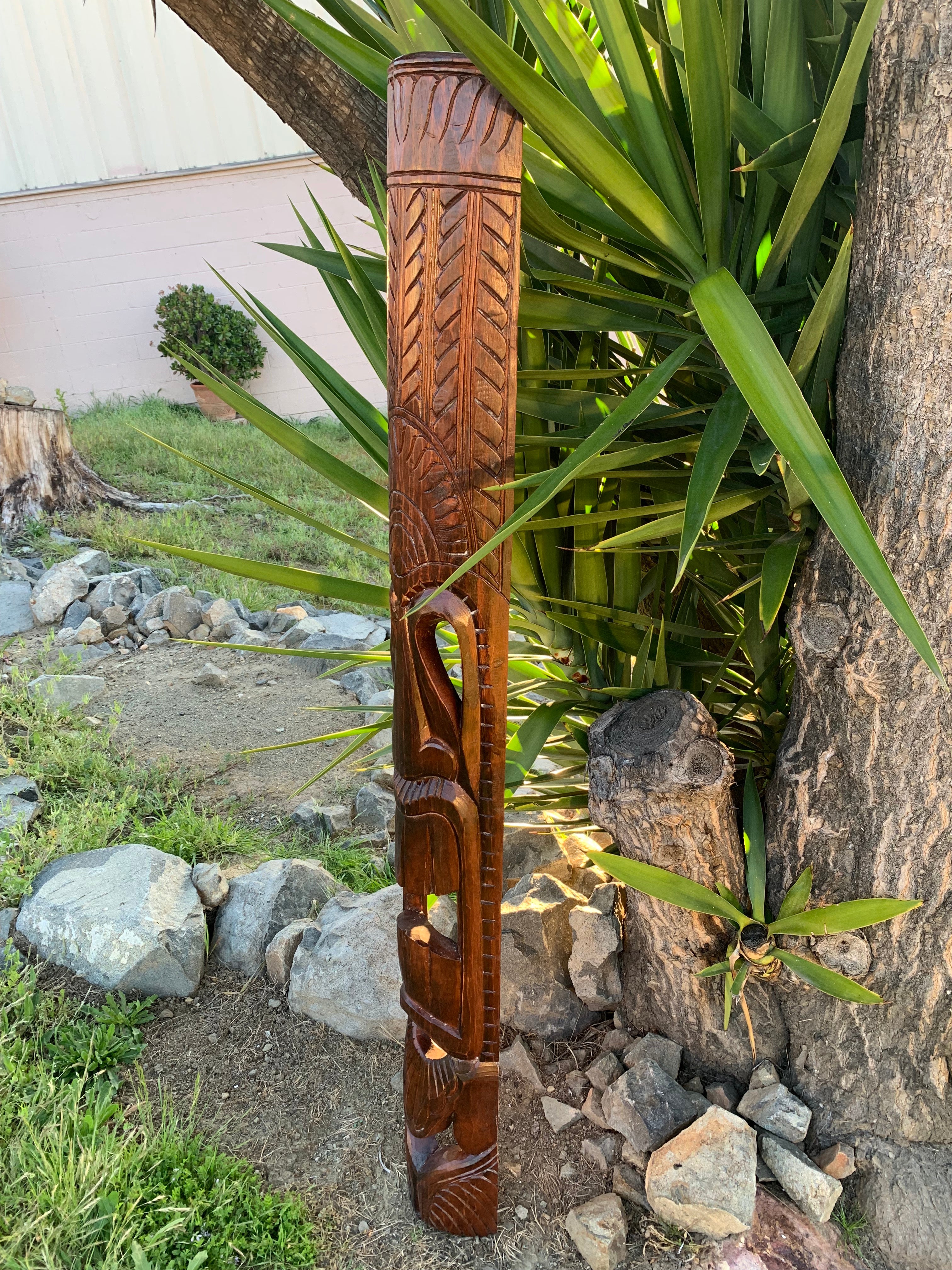 Hand-carved wooden post with tribal geometric patterns standing in a garden beside rocks and greenery.