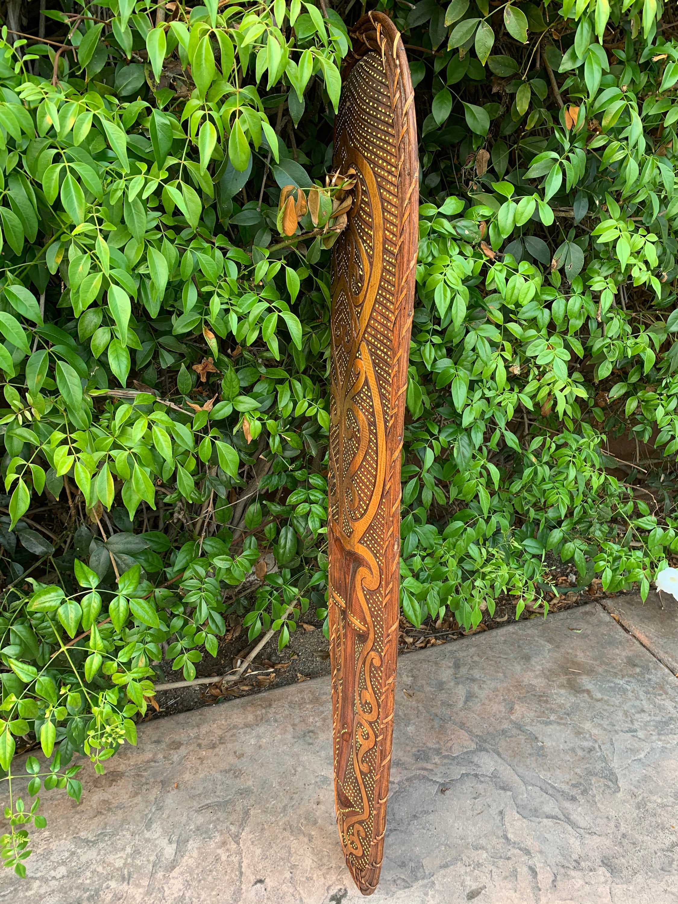 Hand-carved wooden staff with tribal patterns leaning against a green leafy wall outdoors.