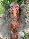 Hand-carved wooden tiki mask sculpture mounted on a tree trunk outdoors.
