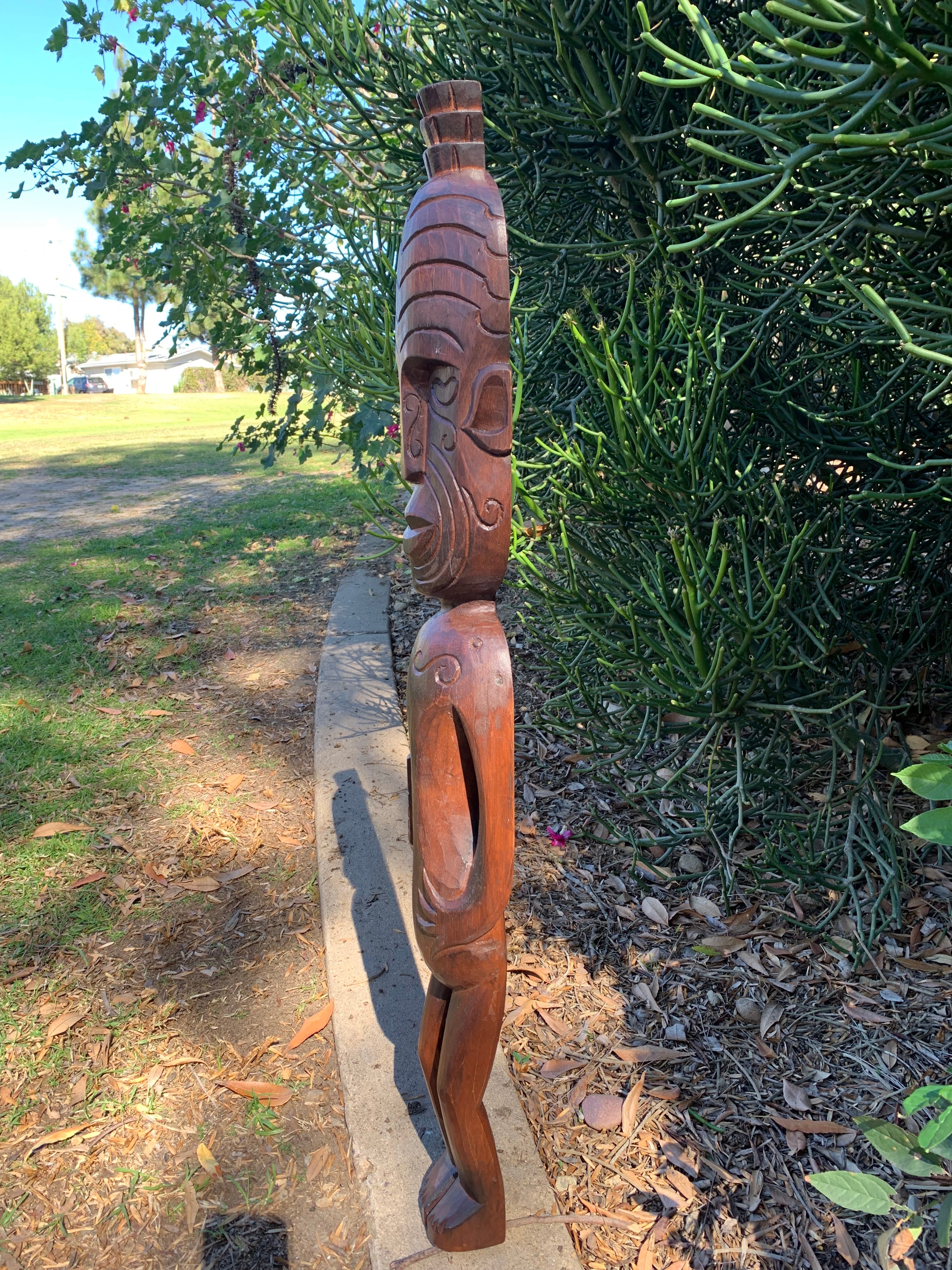 Hand-carved wooden tiki statue outdoors beside a green shrub in sunlight