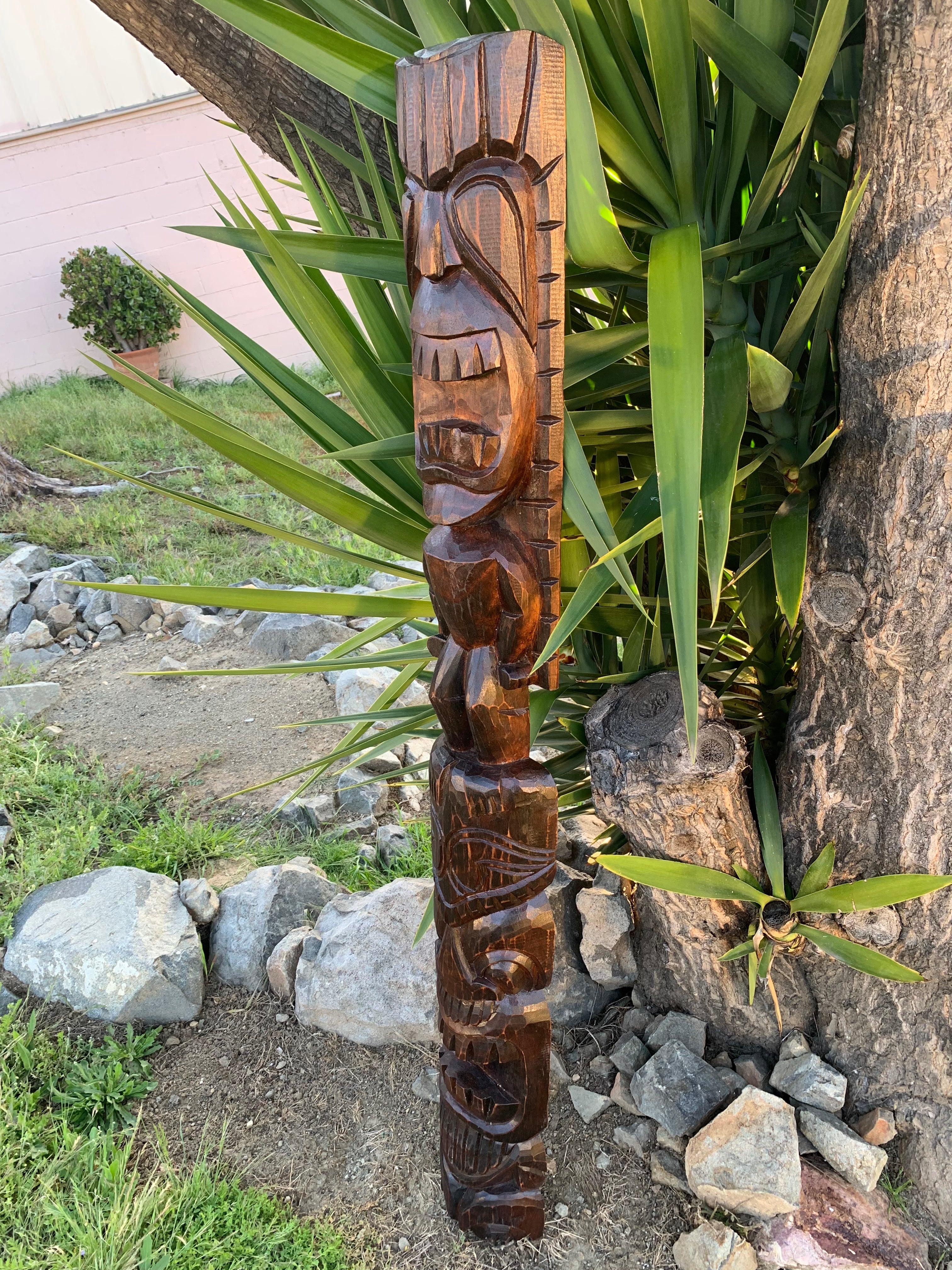 Hand-carved wooden tiki totem pole outdoors among plants and rocks.