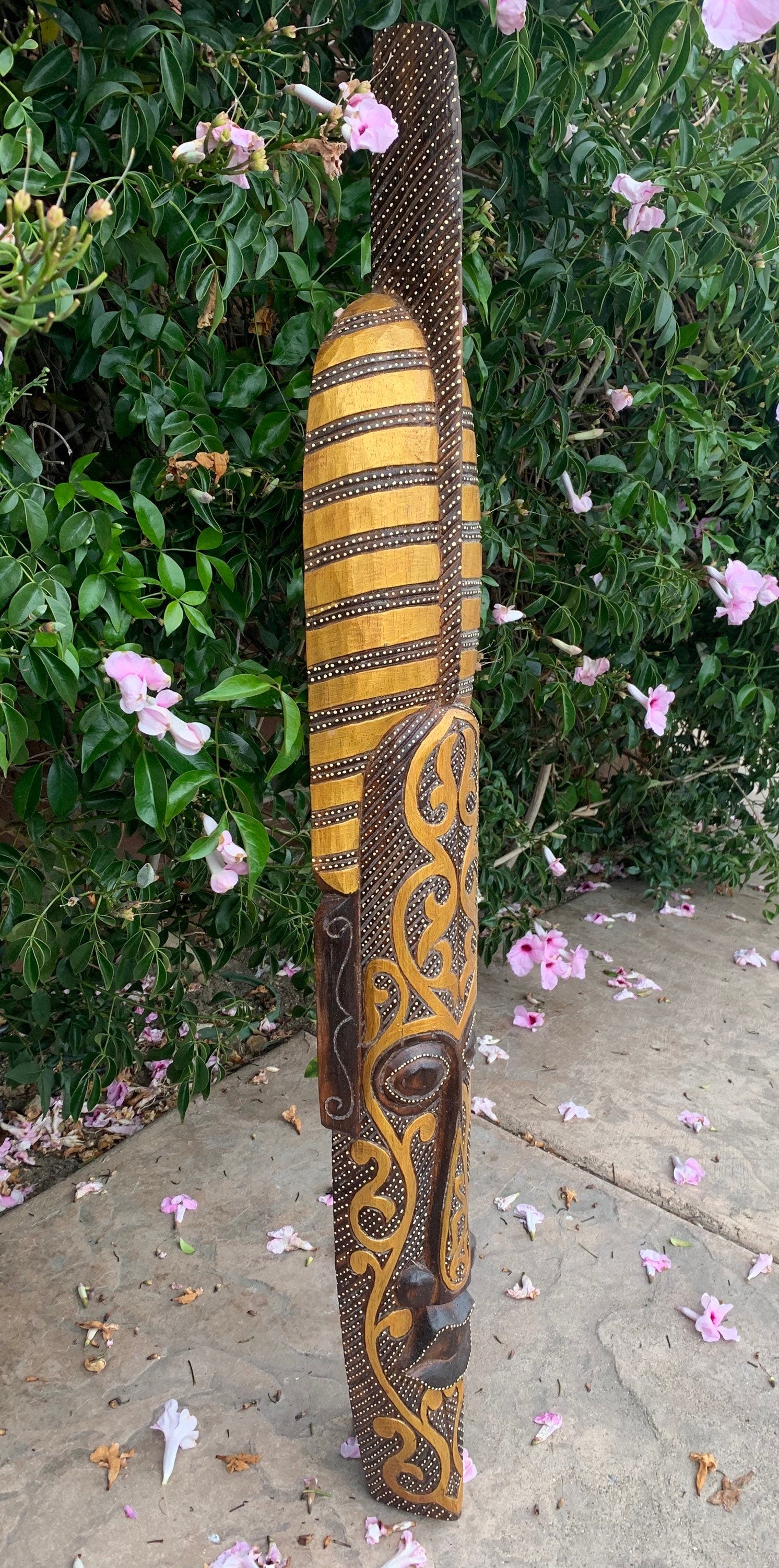 Hand-carved wooden tiki totem pole with gold and brown patterns against green shrubbery and pink blossoms.