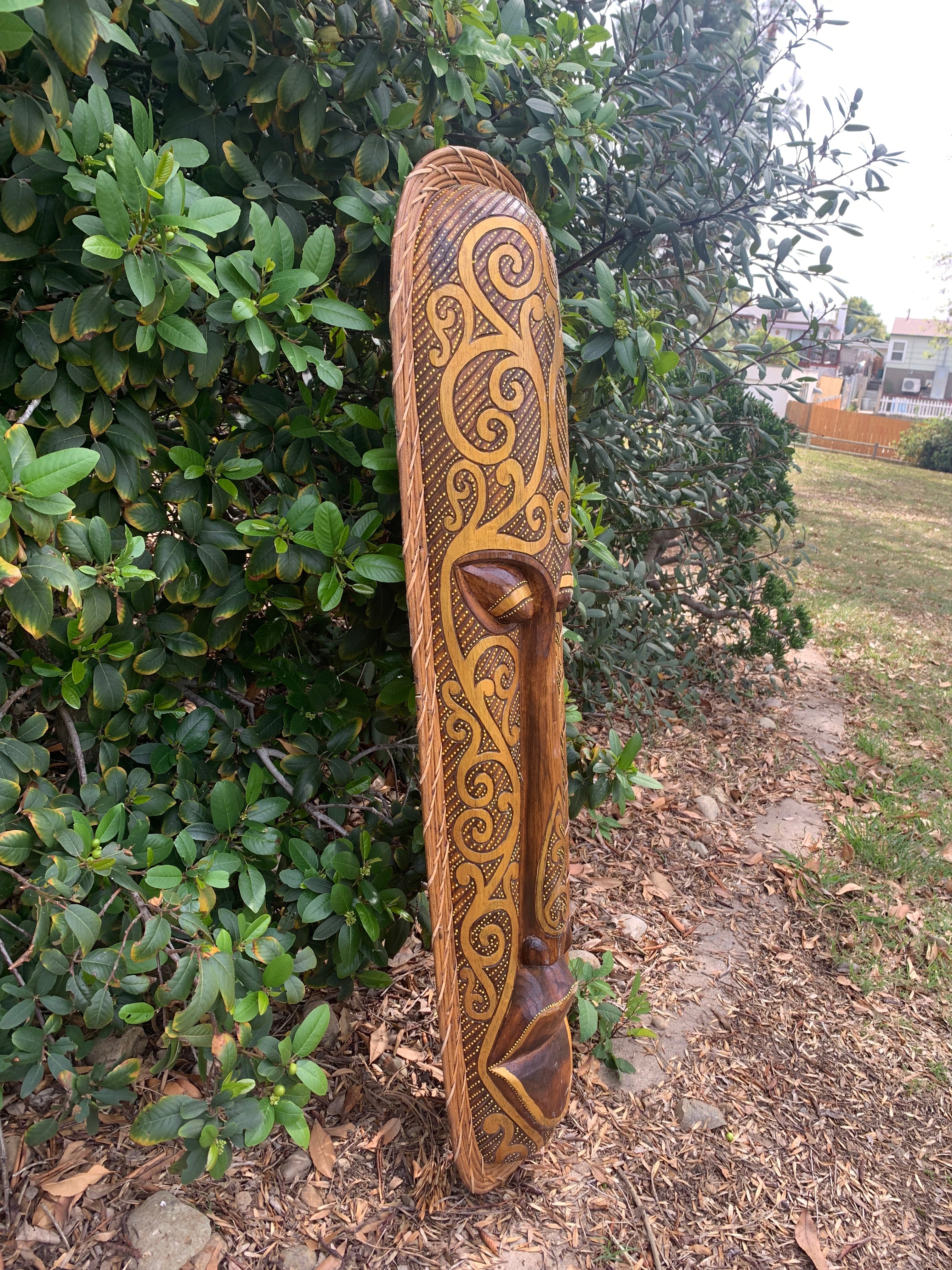 Tall carved wooden totem pole with gold swirling patterns leaning against a hedge.