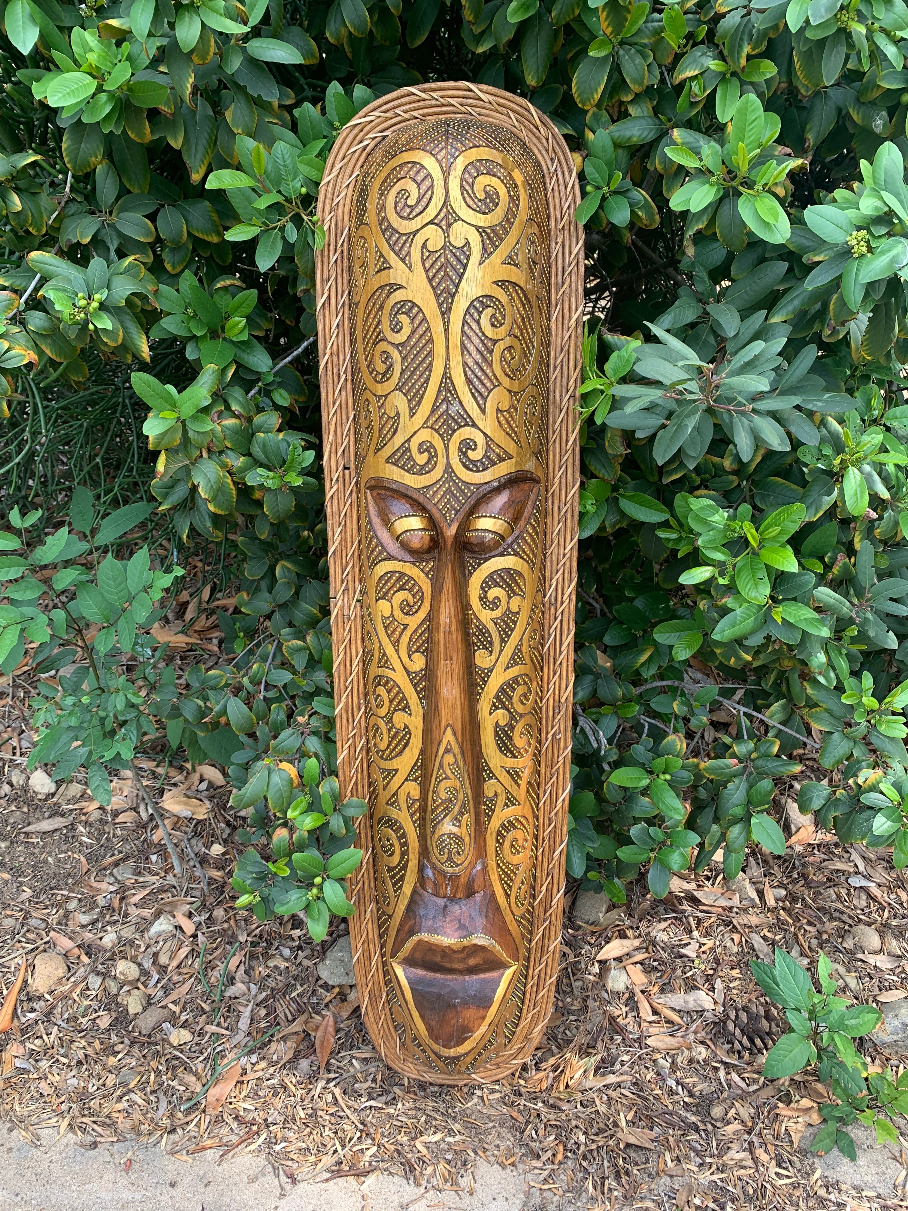 Hand-carved wooden tribal mask with swirling patterns, outdoors among green foliage.