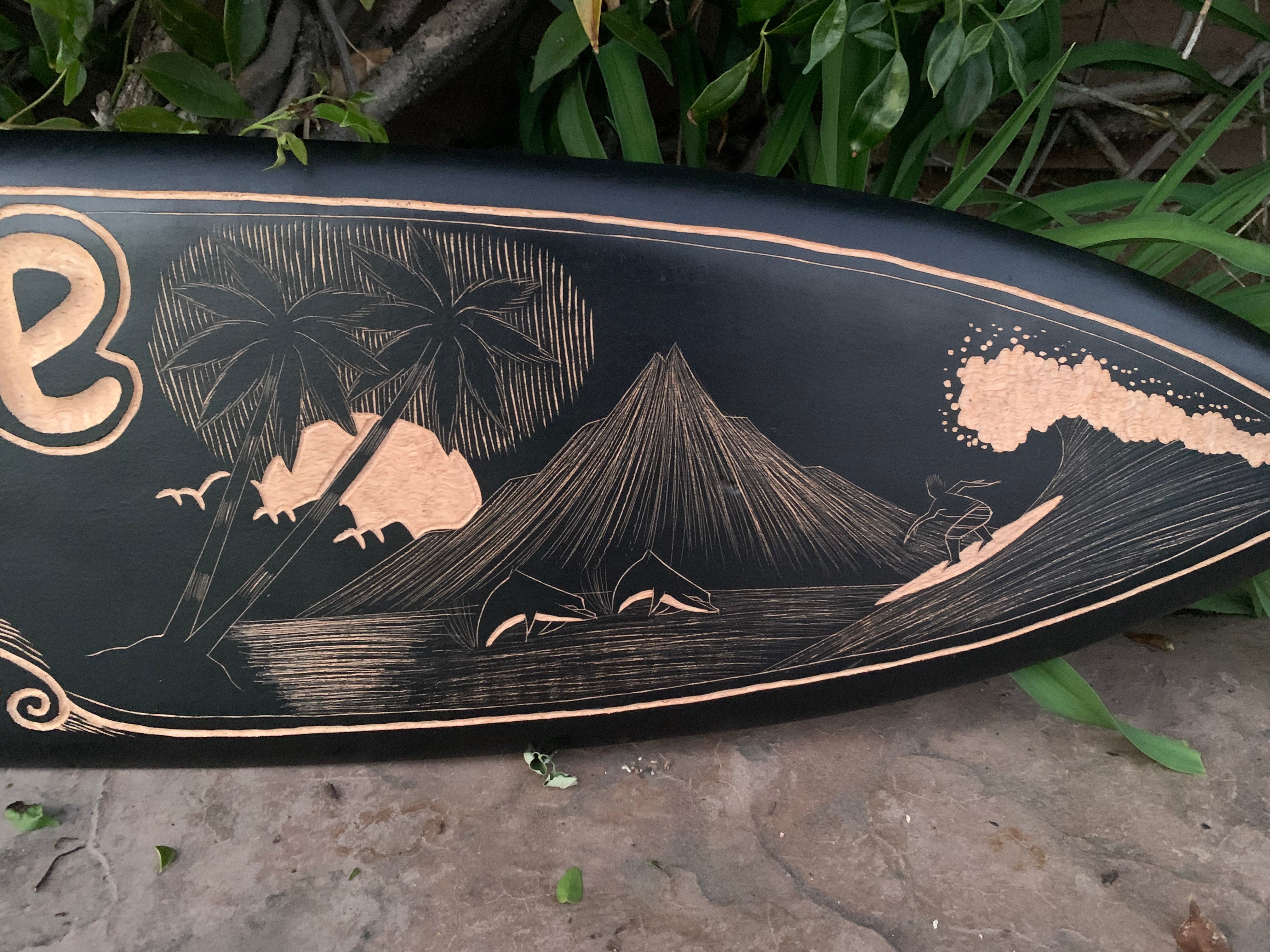 Black surfboard with gold-etched Hawaiian scene showing palm trees, a volcano, and ocean waves