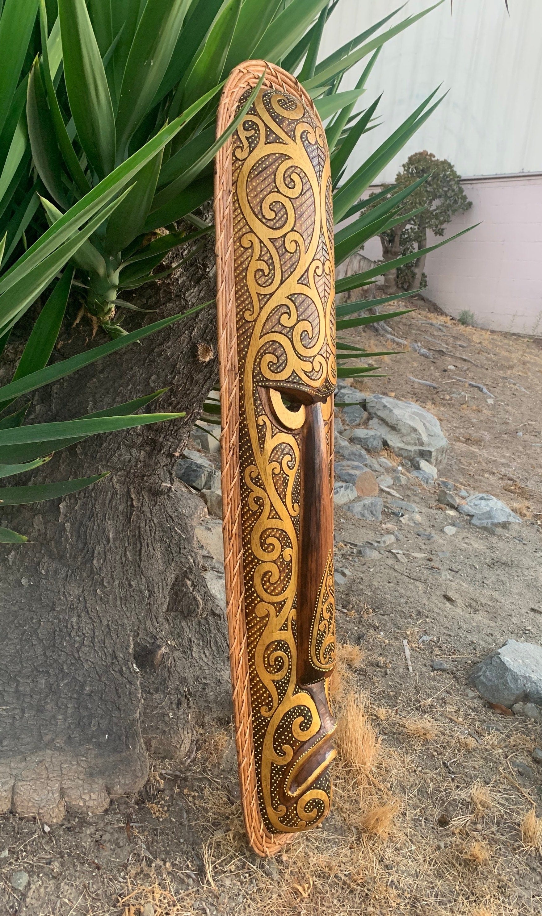 Tall carved wooden object with intricate gold-brown swirl patterns and a woven rope edge leaning against a tree.
