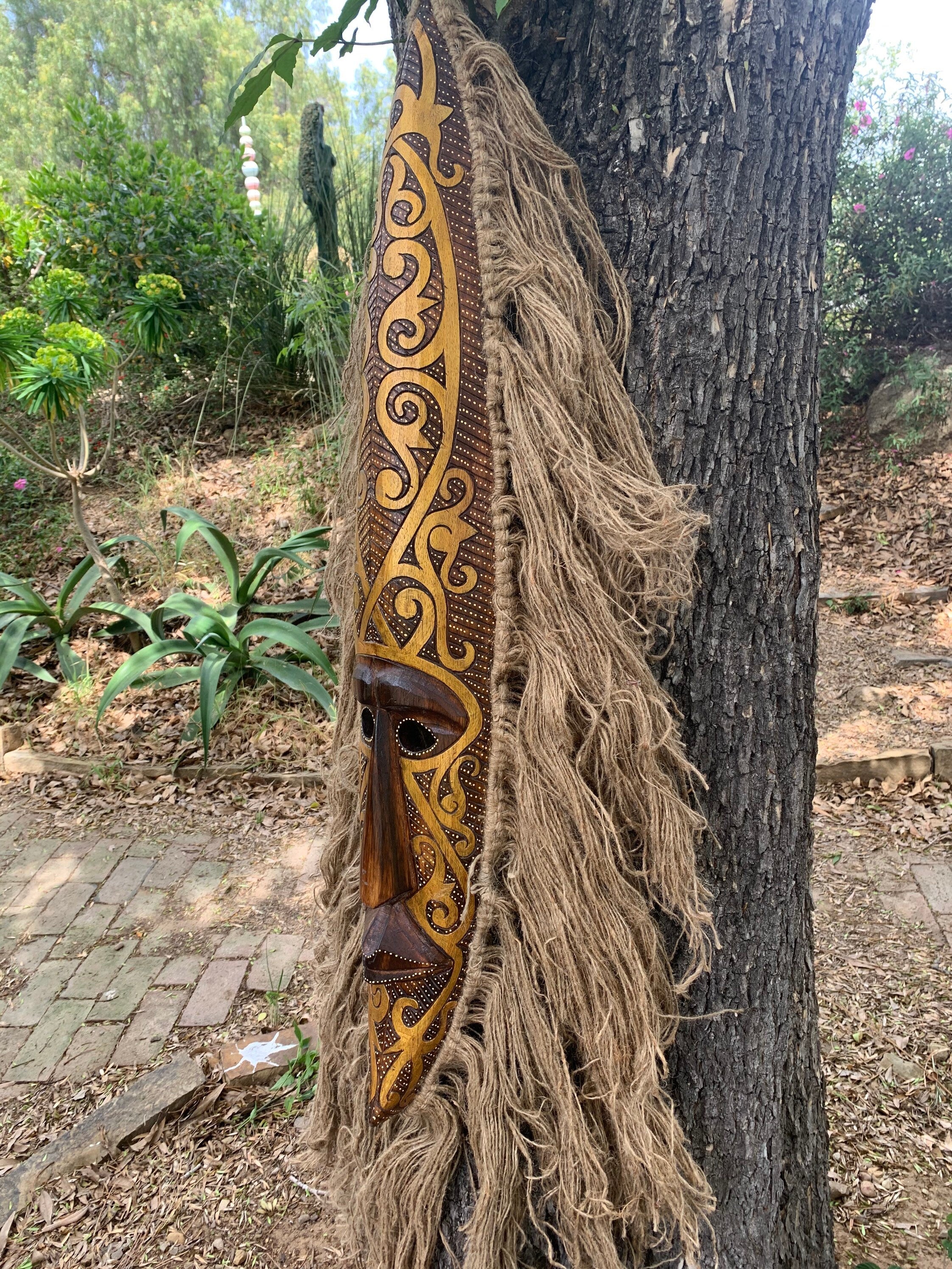 Intricately carved wooden mask with gold patterns and long natural-fiber fringe hanging on a tree.