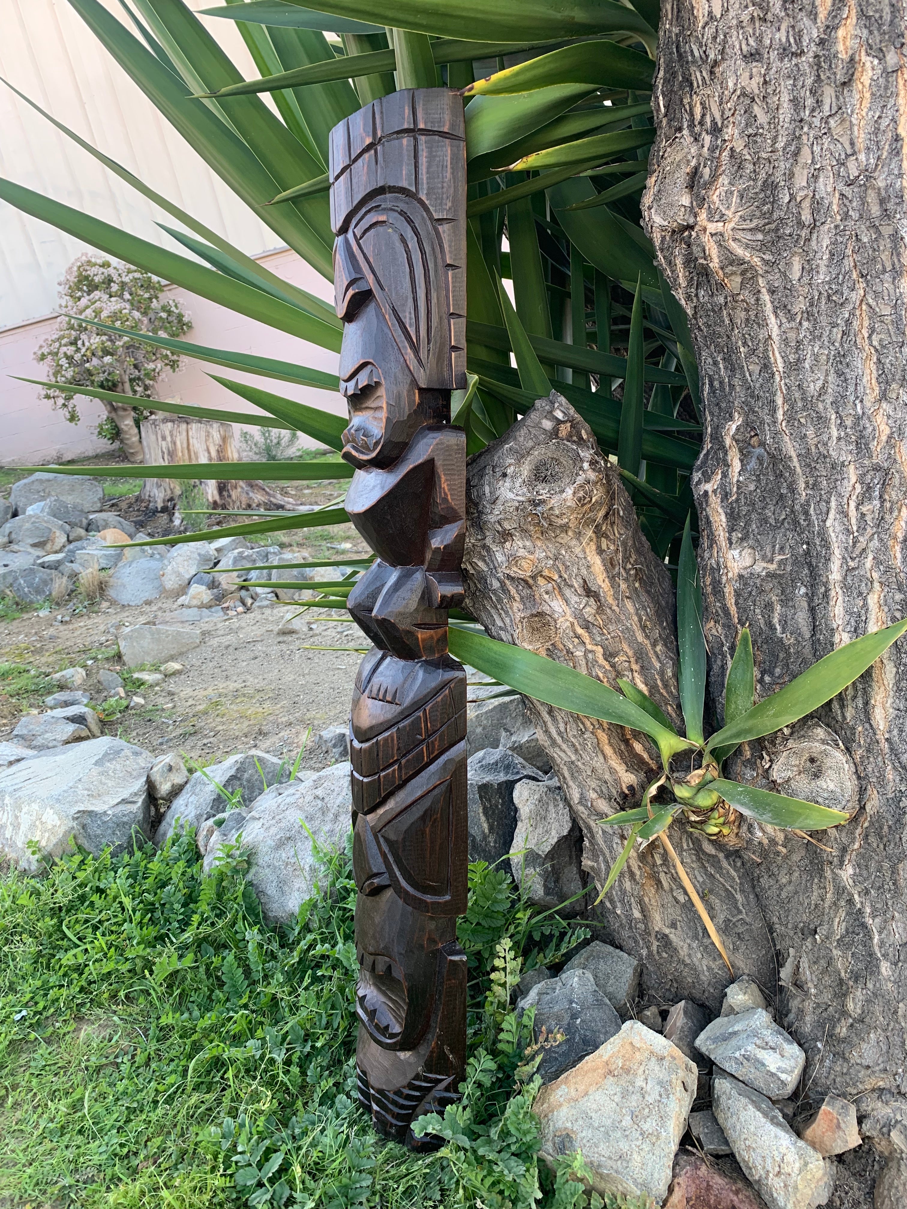 Intricately carved dark wood totem pole leaning against a tree in a garden.