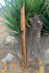 Long carved wooden staff with swirling patterns leaning against a tree in a dry landscape.