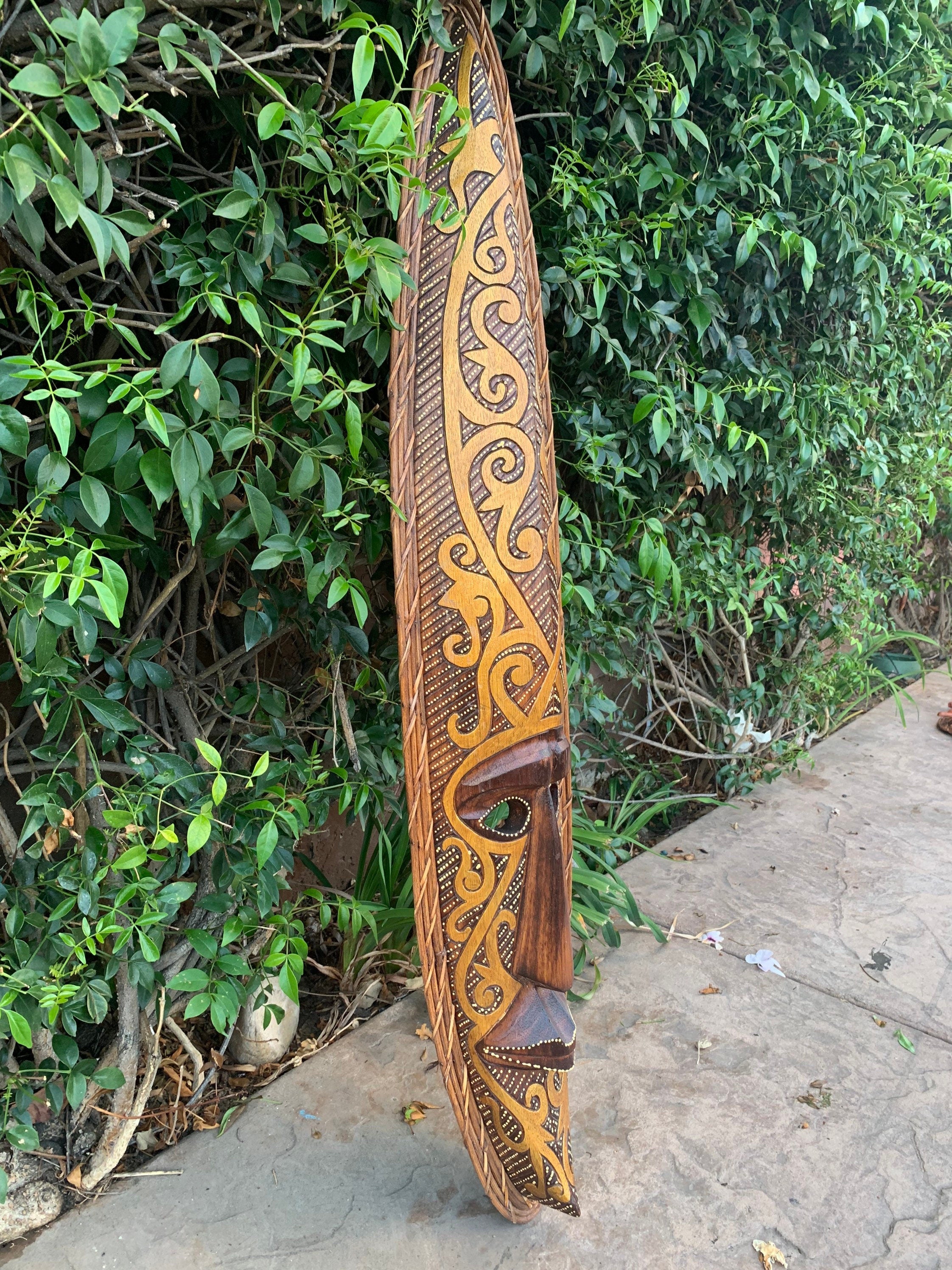 Intricately carved wooden tiki with swirling spiral patterns leaning against a green hedge.