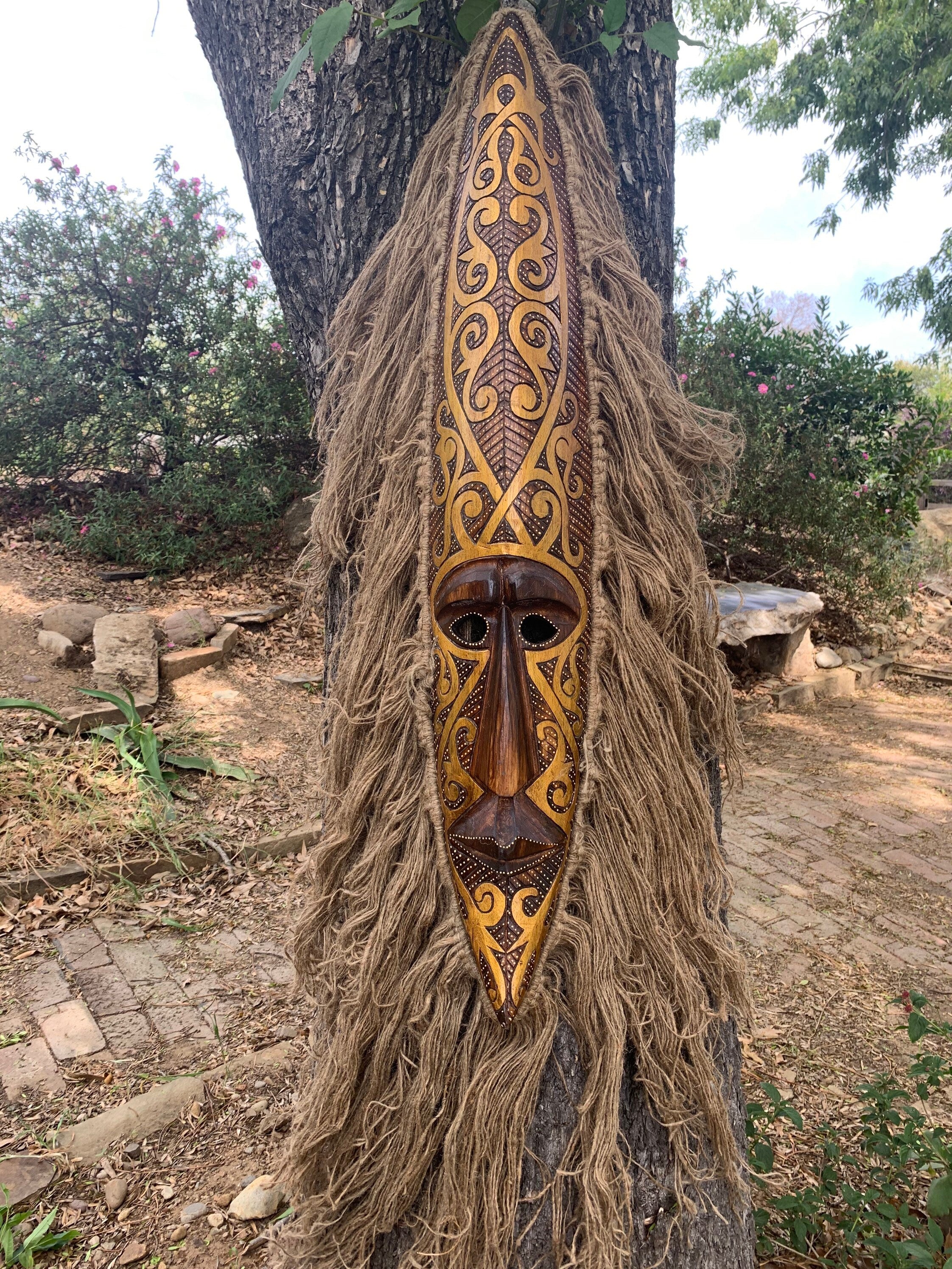 Intricately carved wooden tribal mask with gold swirls and fiber fringe hanging on a tree outdoors