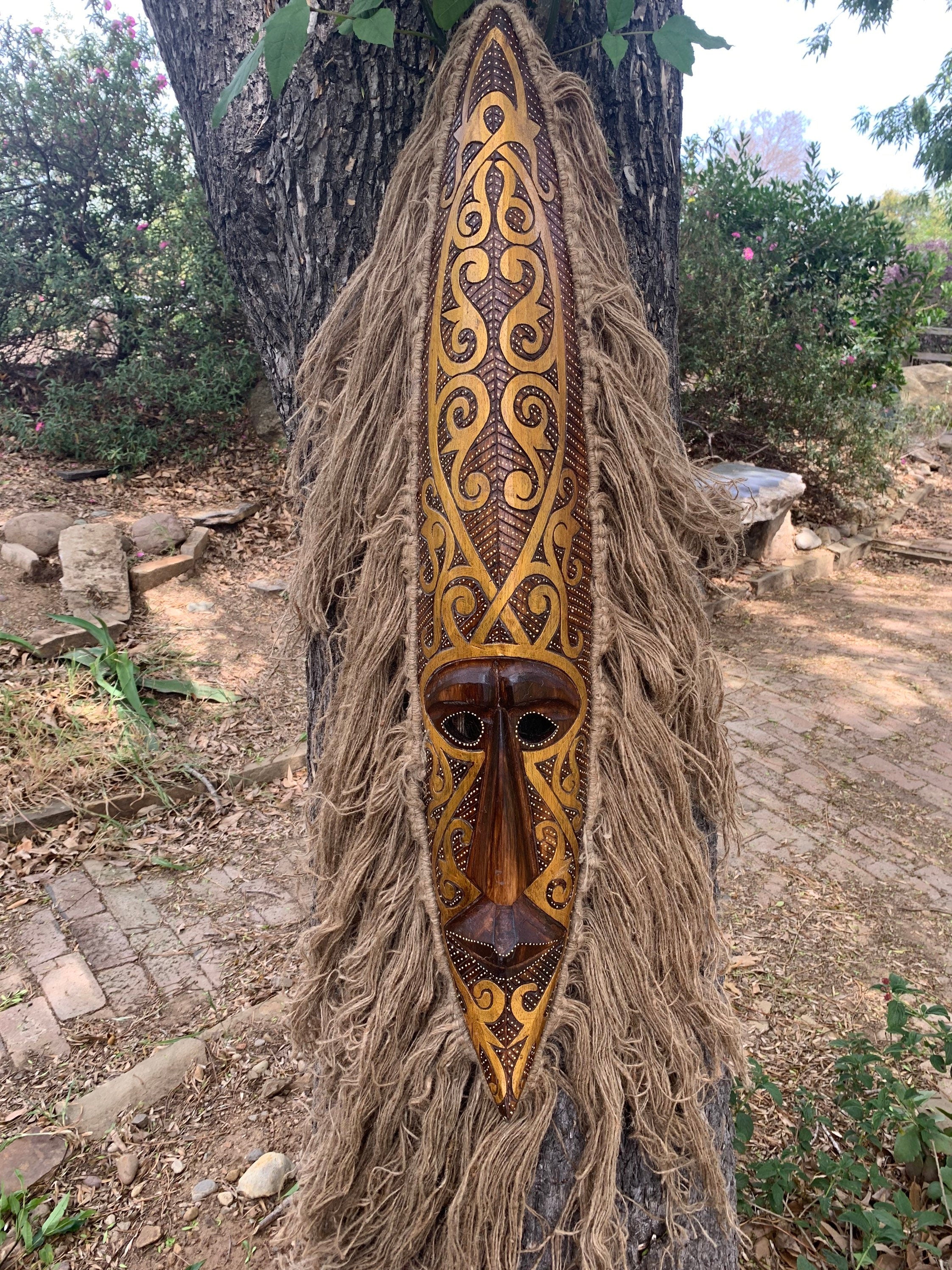 Intricately carved wooden tribal mask with gold patterns and fiber fringe hanging on a tree.