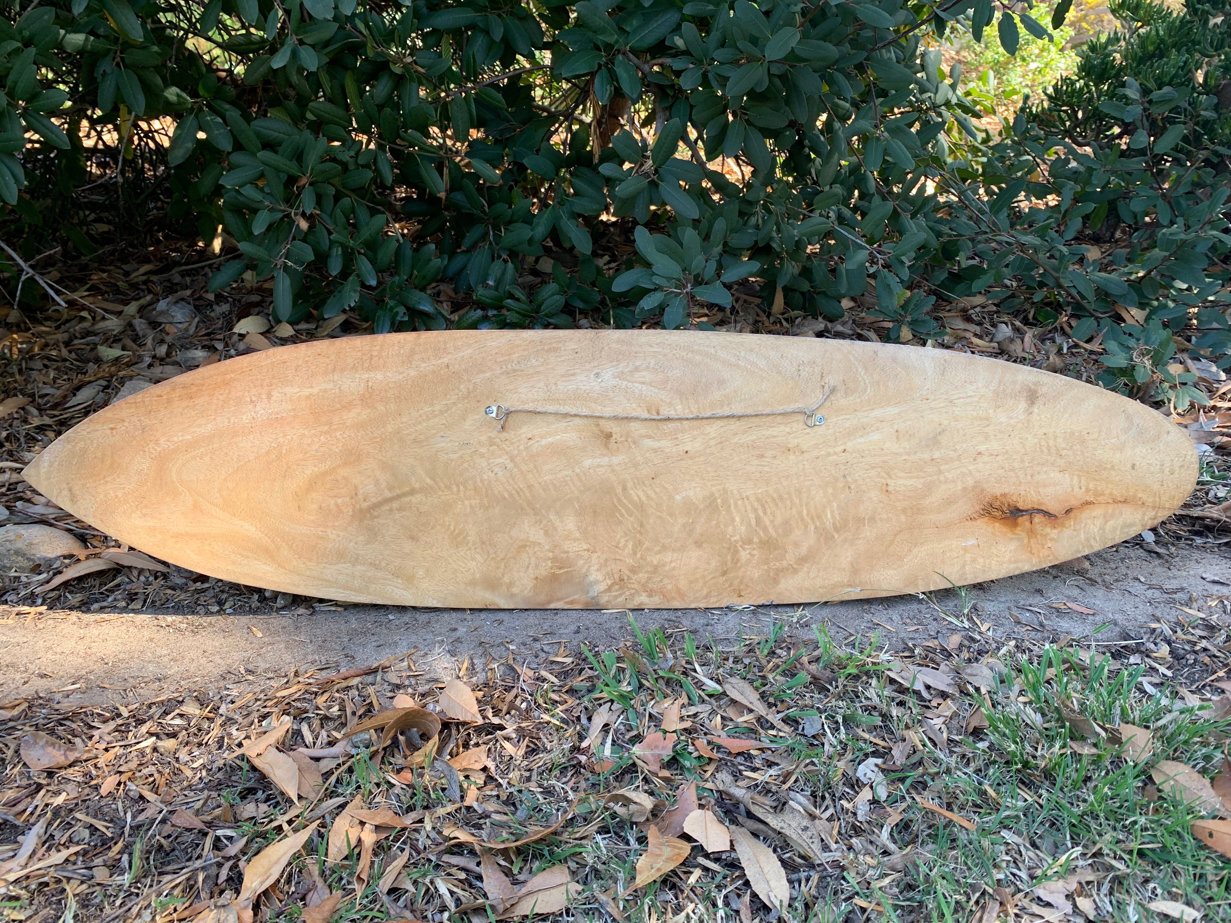 long natural wooden surfboard resting on the ground beside green bushes