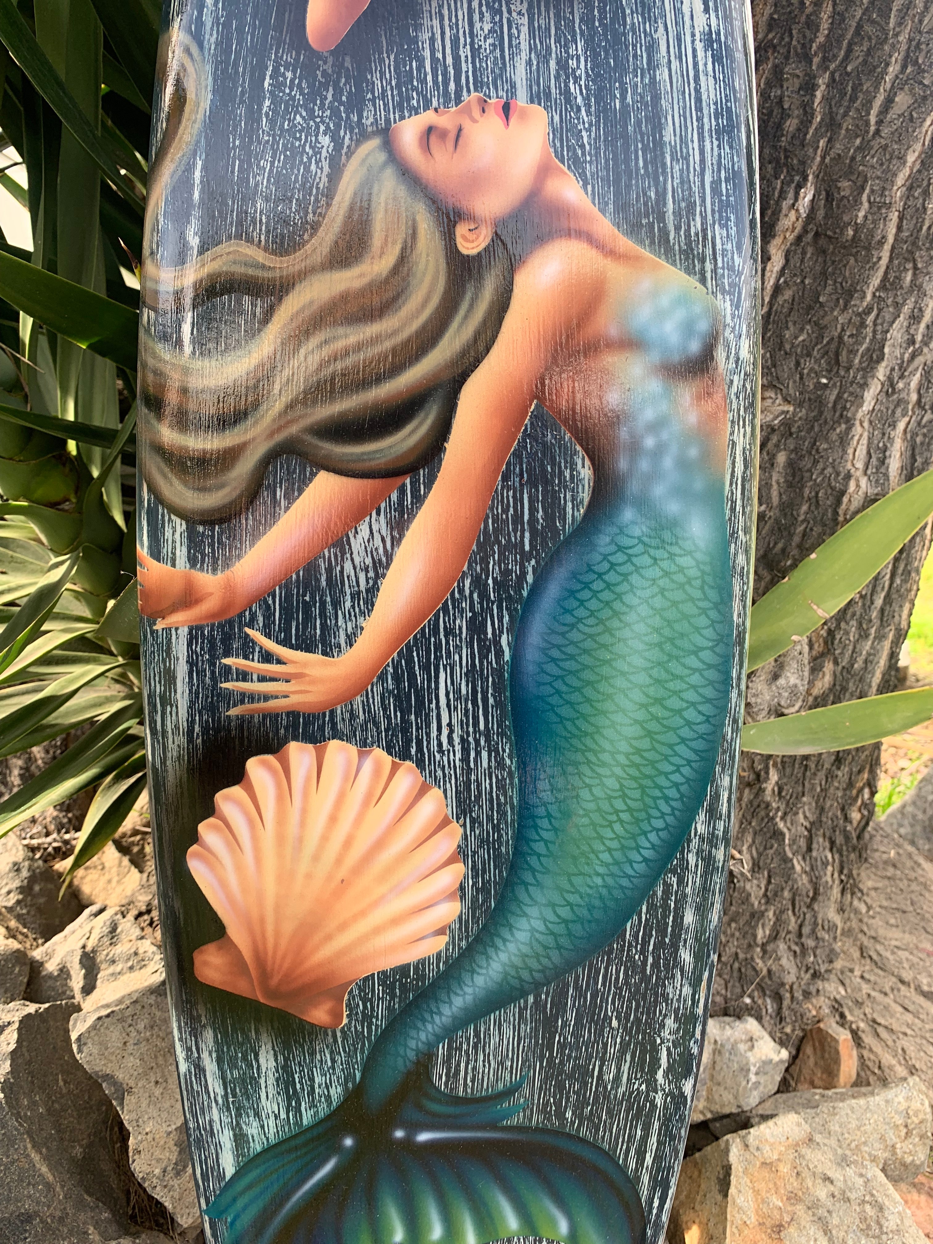 Mermaid illustration with flowing blonde hair and green-scale tail on a weathered board beside a seashell.