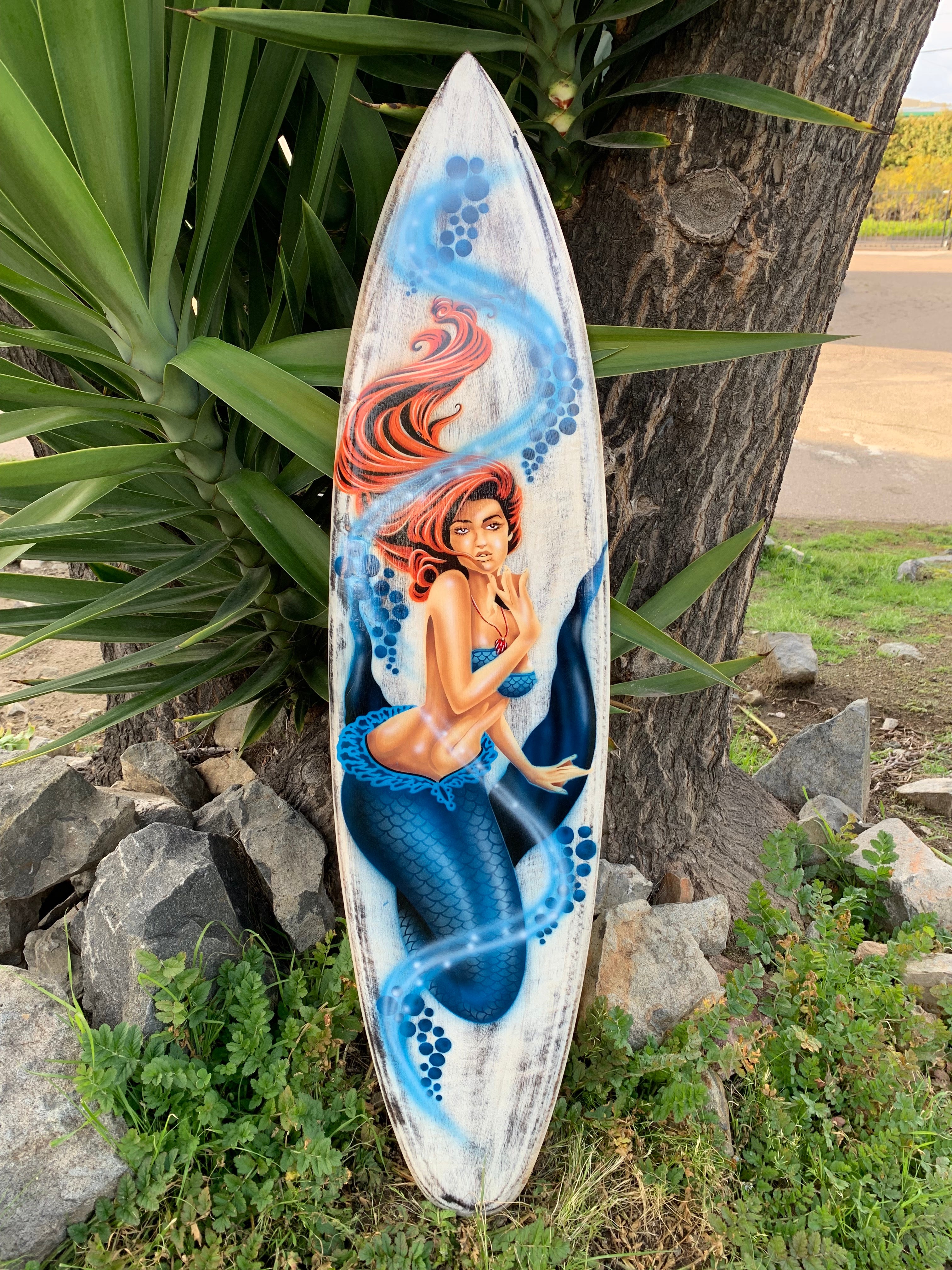 Mermaid illustration on a weathered surfboard leaning against a tree