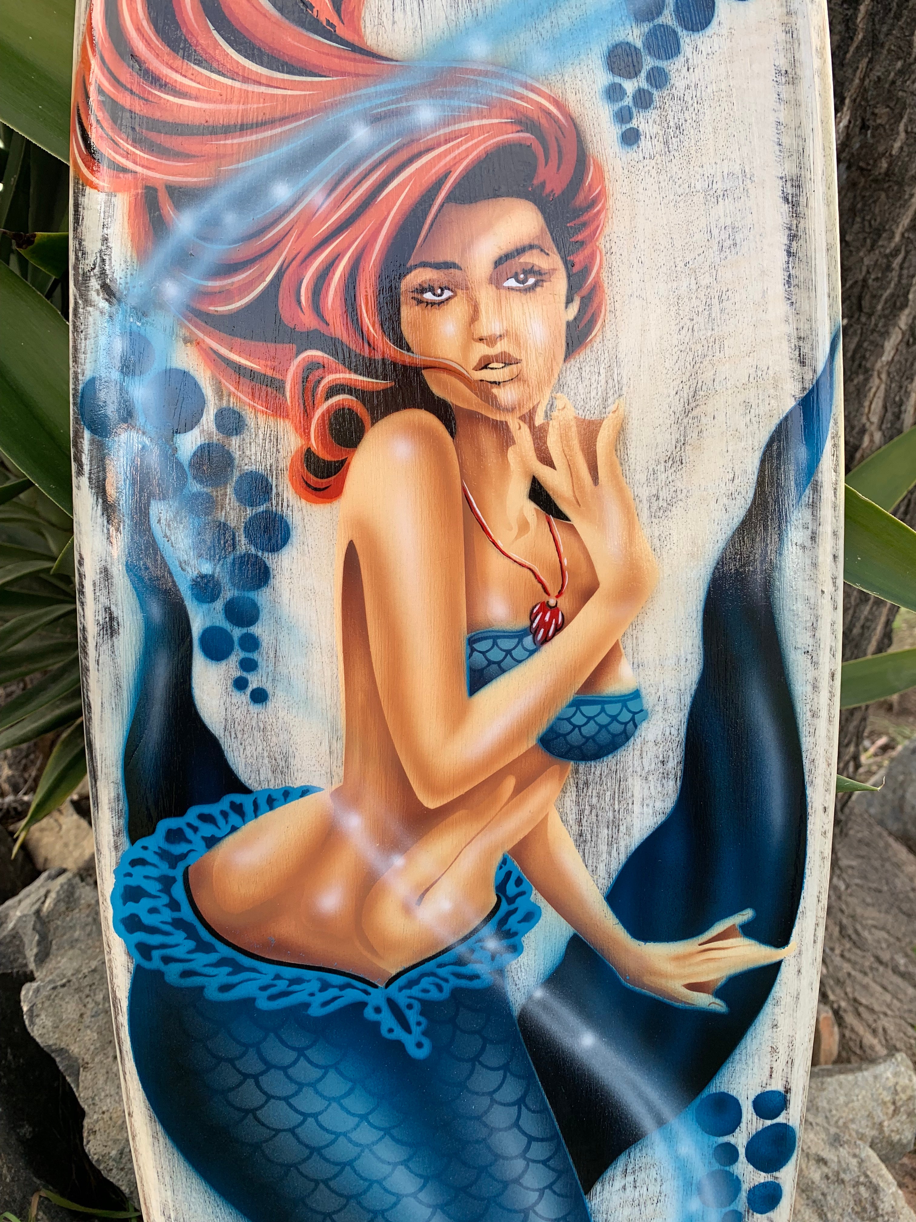Vibrant mermaid illustration with red hair and blue tail painted on a weathered wooden board.