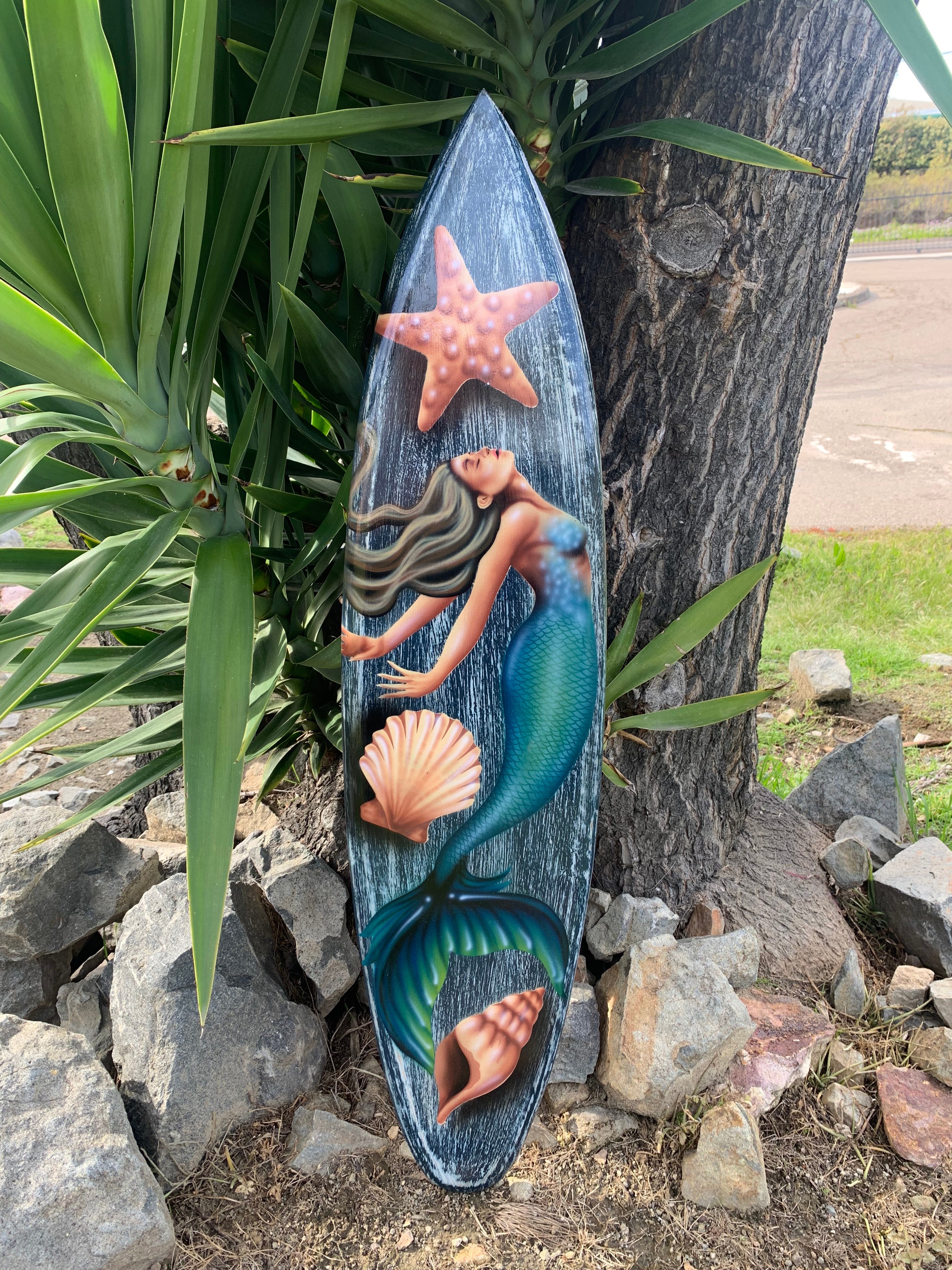 Mermaid-themed decorative surfboard with starfish and seashells leaning by a tree.