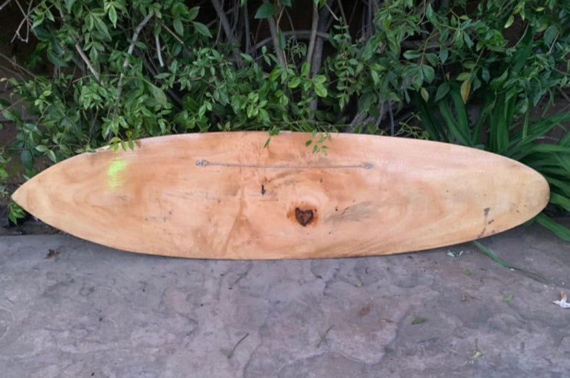 Natural wood longboard surfboard resting on concrete with greenery in the background