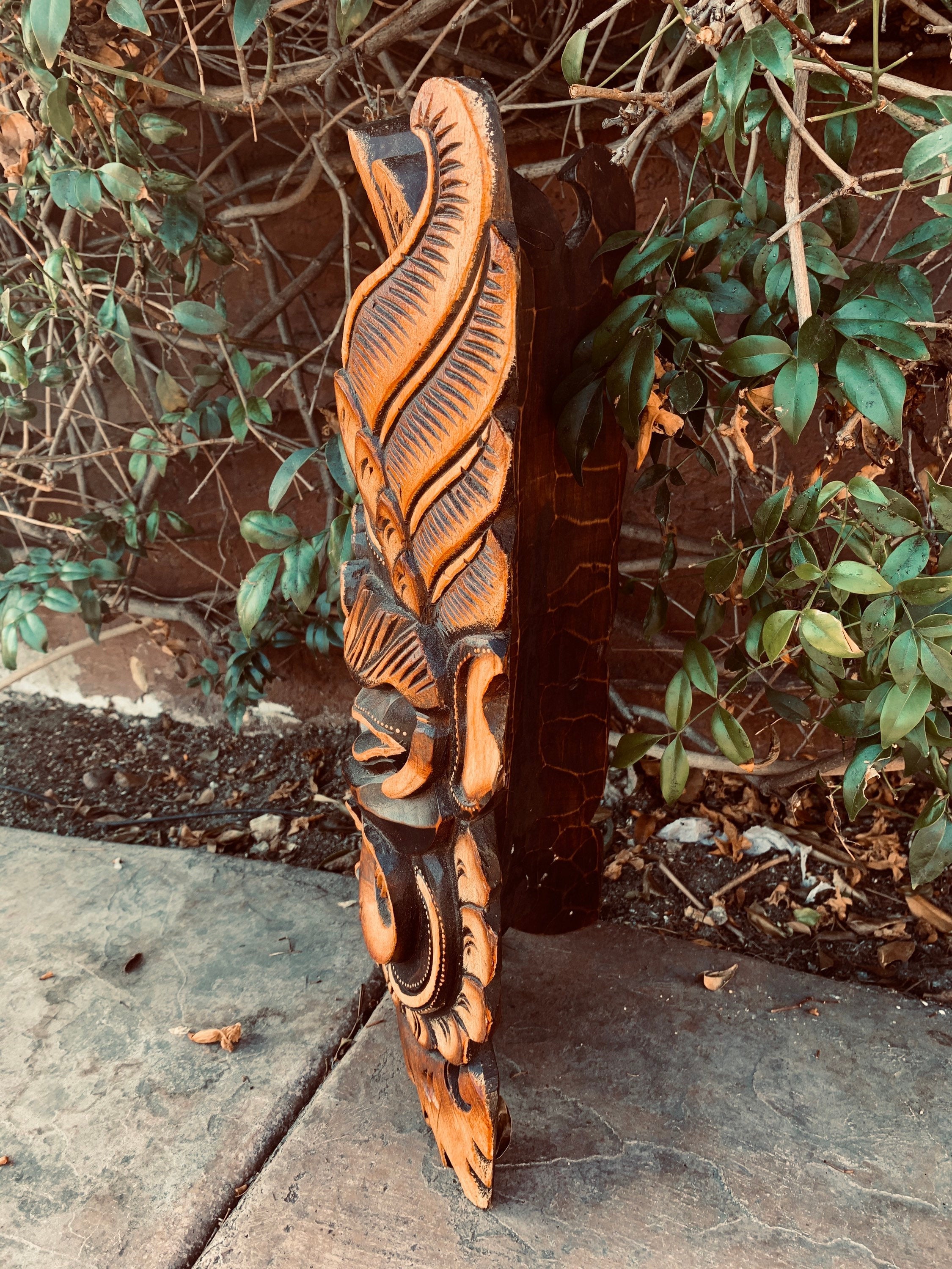 Carved orange and black wooden sculpture with leaf-like patterns leaning against a shrub