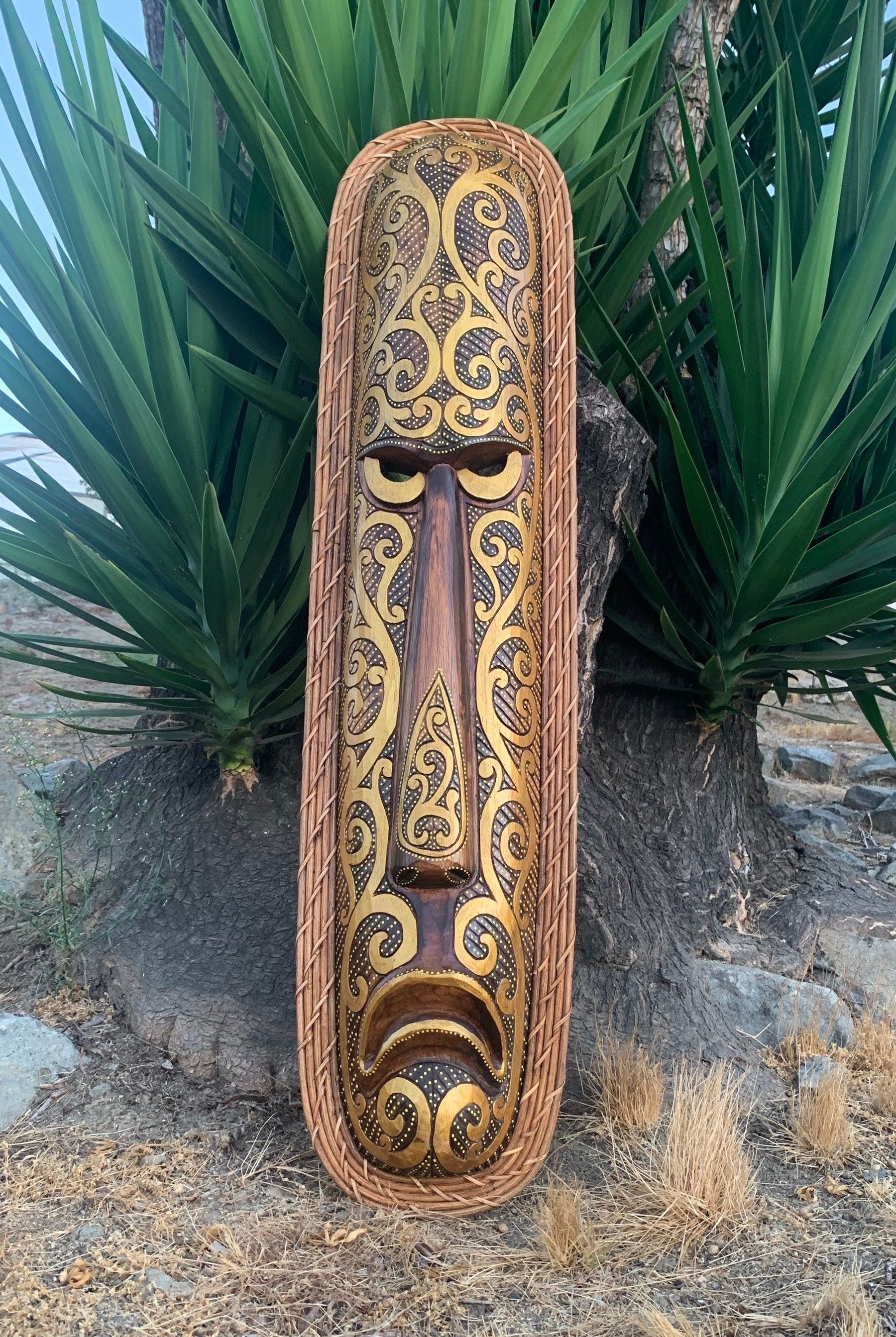 Ornate carved tiki mask with gold scrollwork and braided rope border, displayed outdoors beside green foliage.