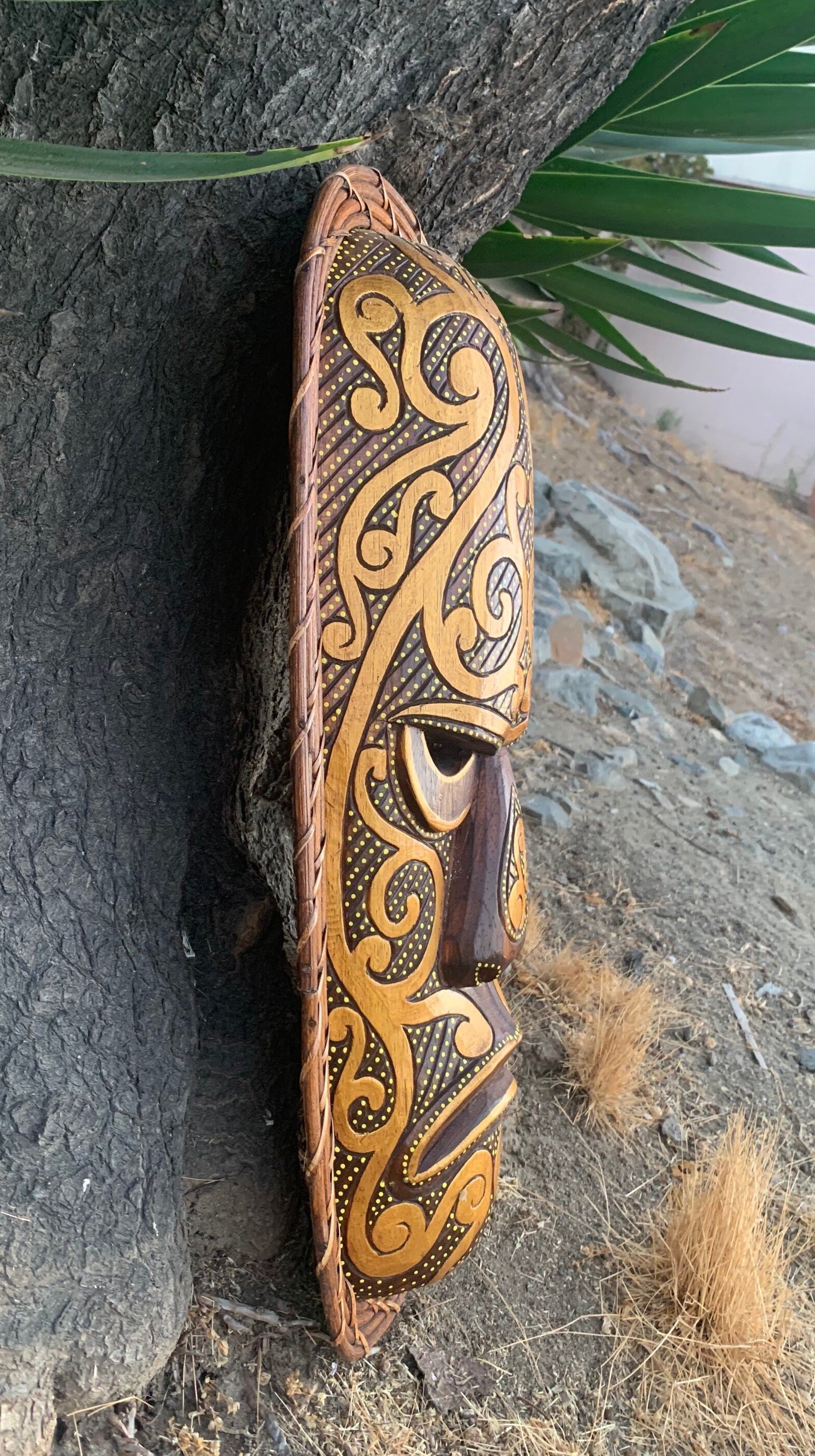Ornate wooden tribal mask with swirling patterns leaning against a tree outdoors