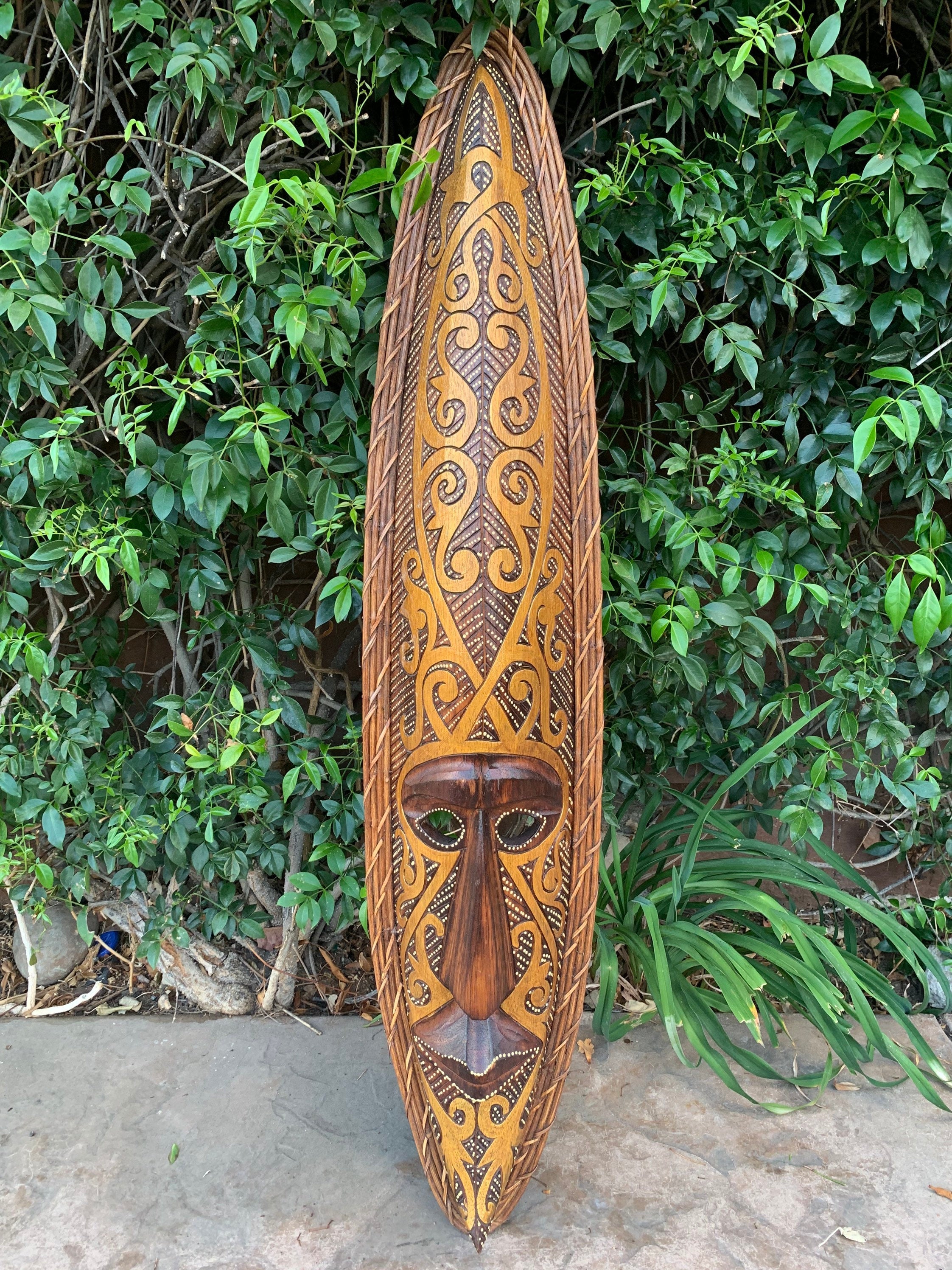 long wooden surfboard with ornate tribal mask carving and scrolling patterns against green foliage