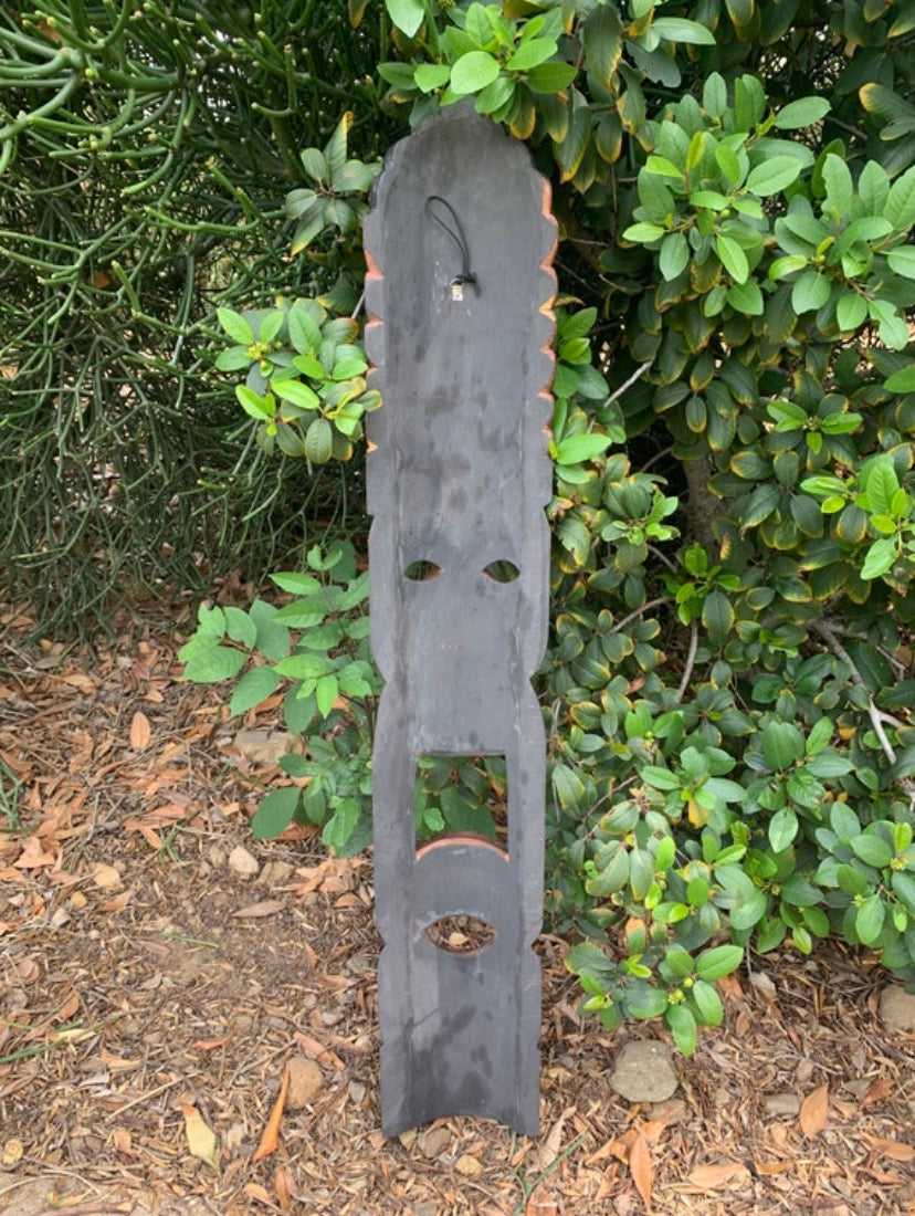 wooden totem-style sculpture with eye holes and mouth cutout leaning against a hedge