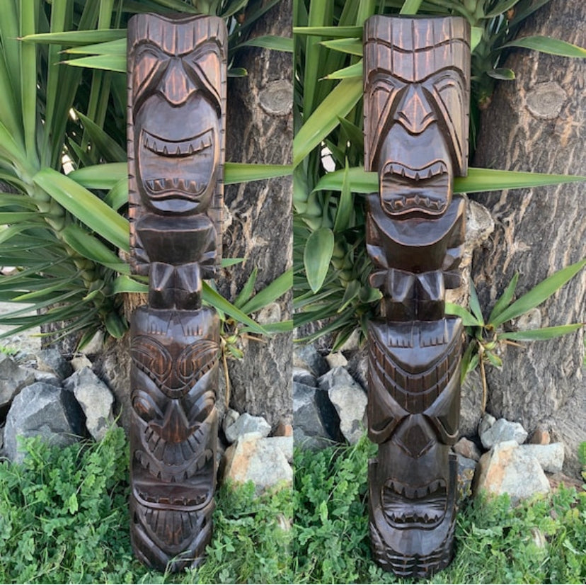 Pair of dark brown carved wooden tiki statues outdoors among plants and stone