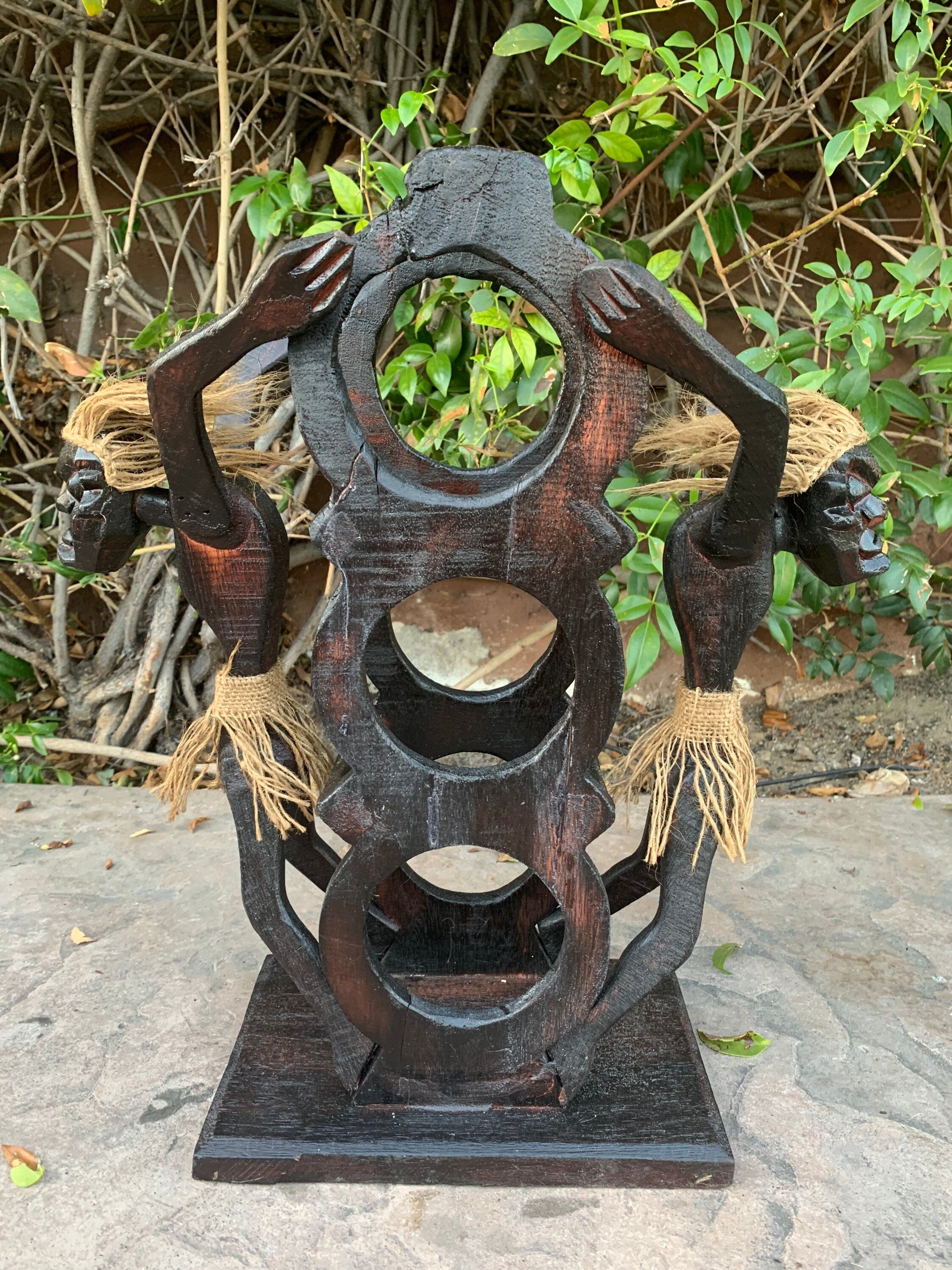 Dark wooden abstract sculpture with circular rings and rope-wrapped arms, outdoors among green plants.