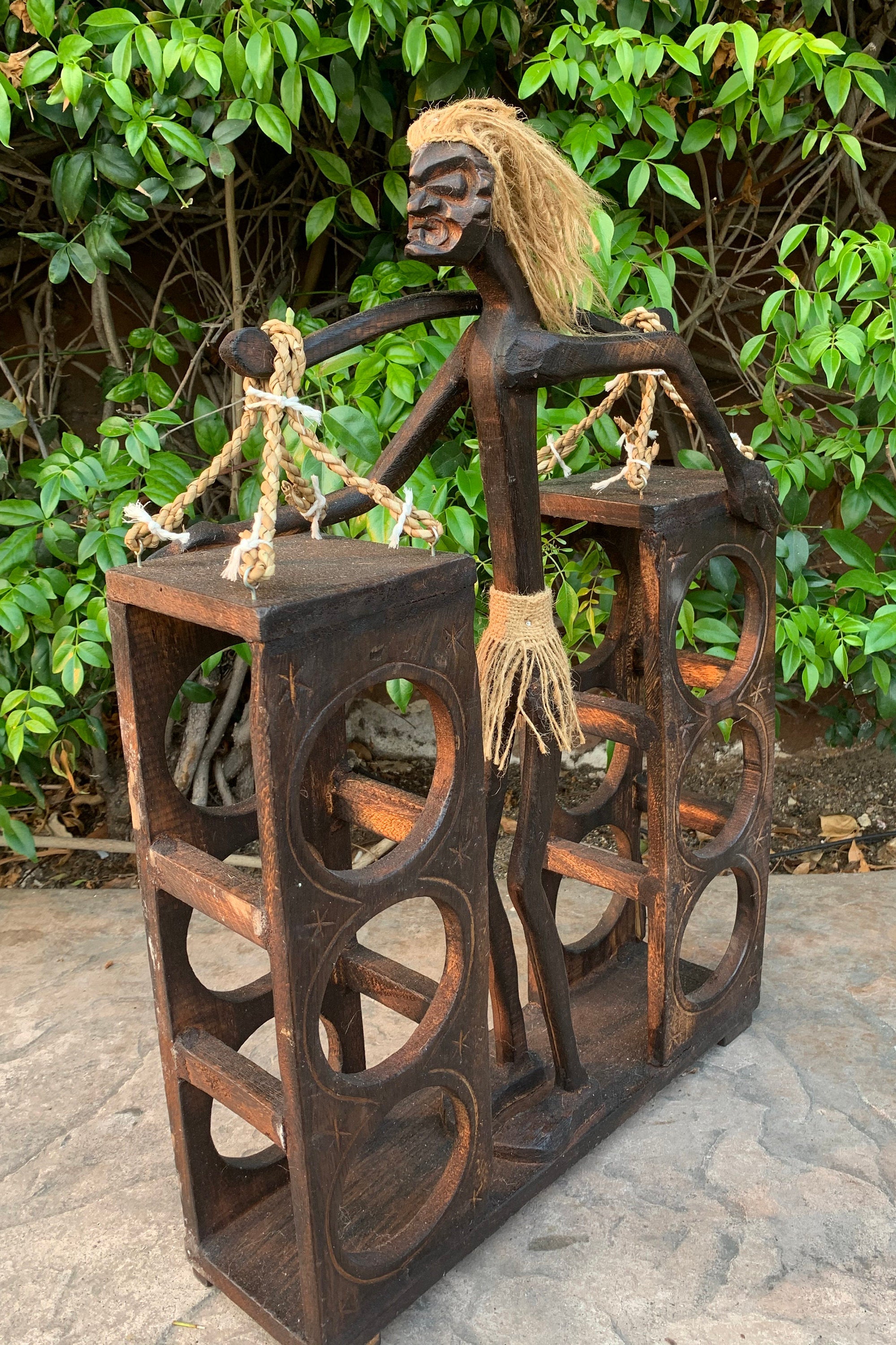 Rustic wooden statue of a standing figure balancing two circular-cut boxes with rope handles.