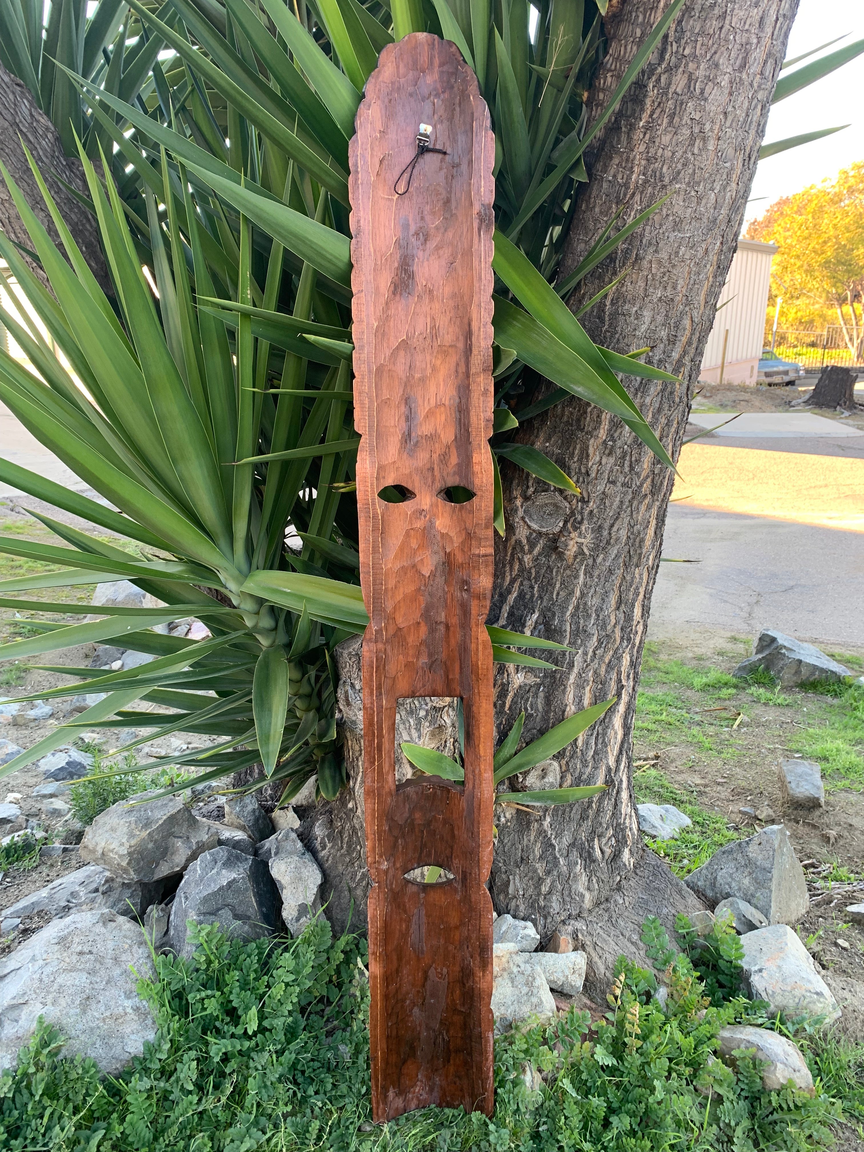 Long rustic wooden wall-hanging with carved holes and a small metal hook, leaning against a tree.