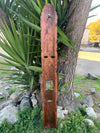 Long rustic wooden wall-hanging with carved holes and a small metal hook, leaning against a tree.