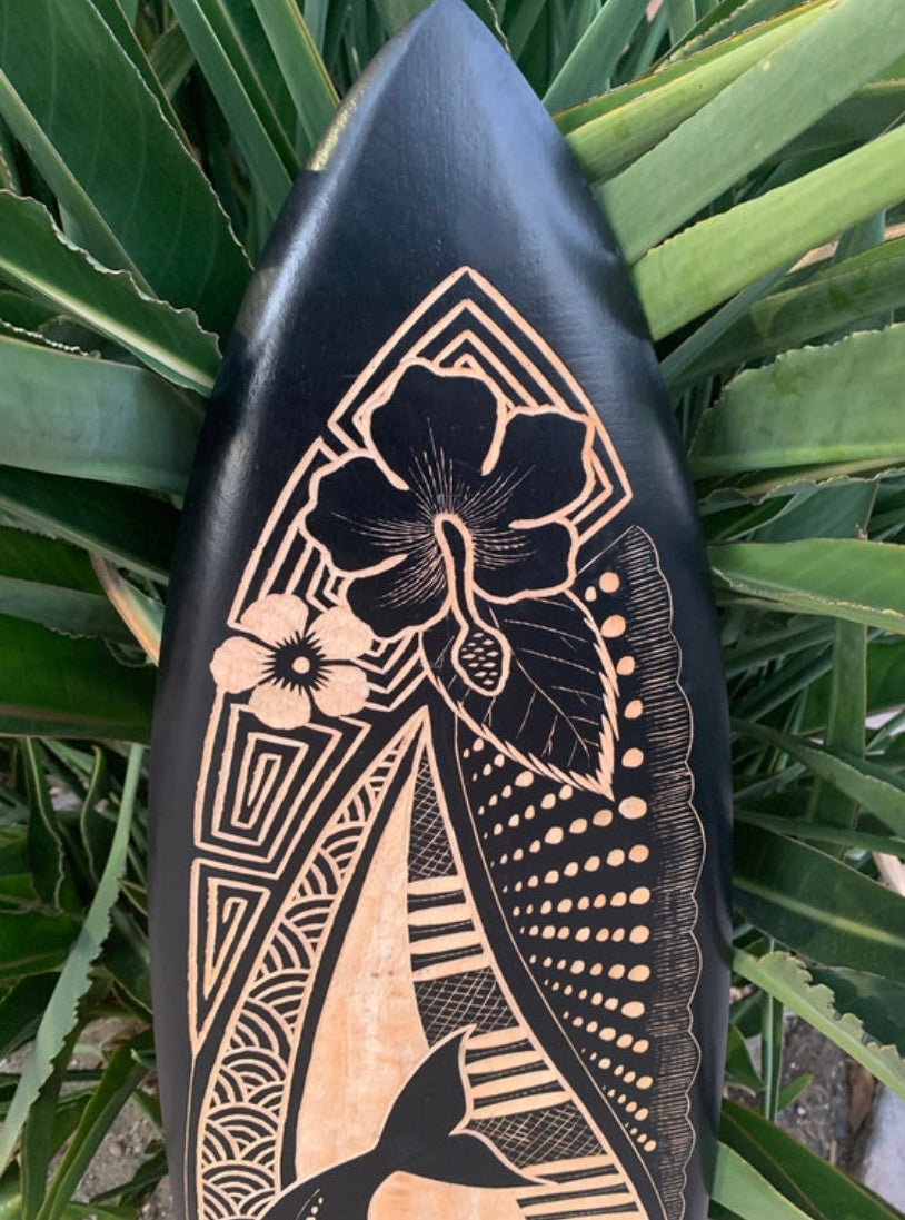 Surfboard with hibiscus flower and geometric wood carvings, black background, among green tropical leaves