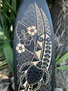 Wood-burned surfboard artwork featuring a sea turtle, hibiscus flowers and stylized ocean waves.