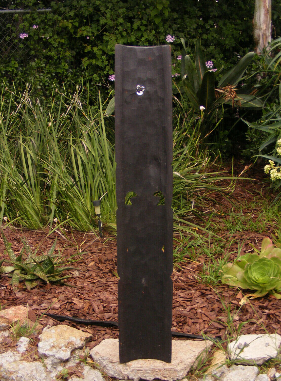 Tall black vertical garden sculpture among mulch and plants