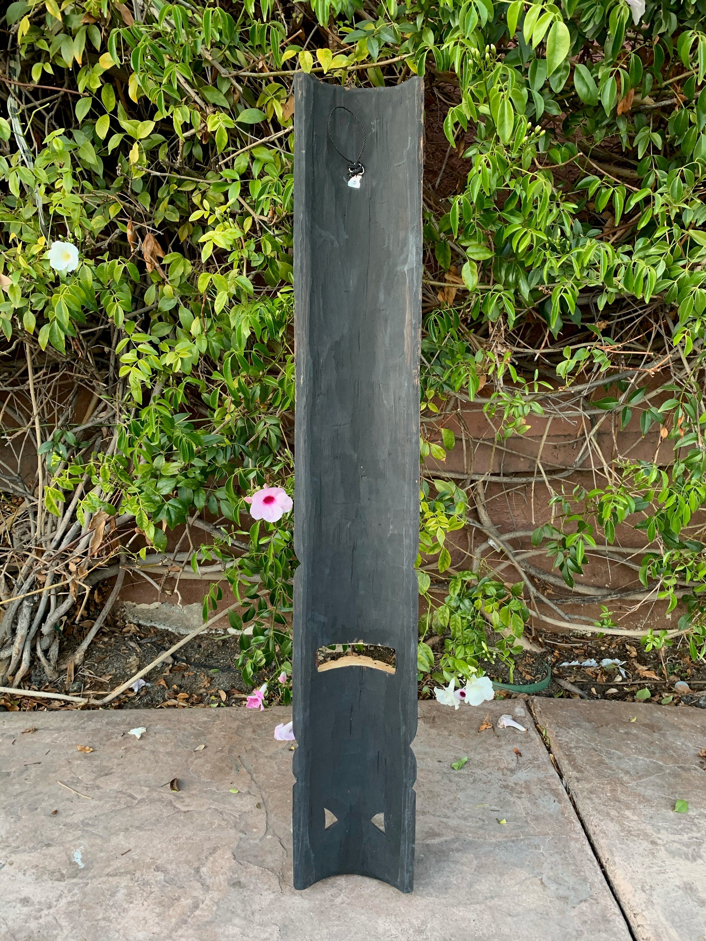 Tall black wooden post with carved cutouts and a small pendant at the top, set against green vines and blooming flowers.