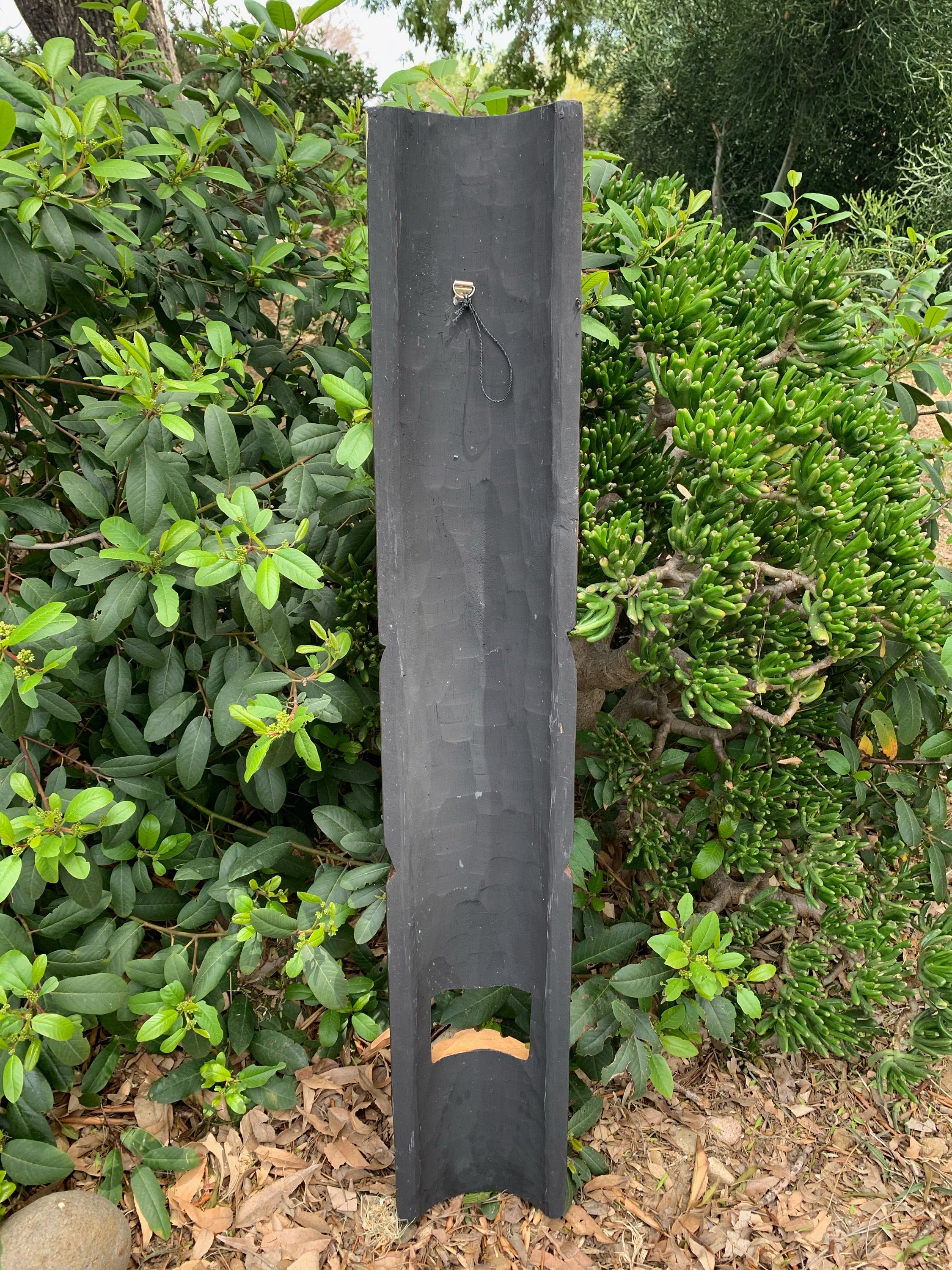 Tall black wooden post with a small metal hook in a garden surrounded by green shrubs.
