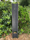 Tall black wooden post with a small metal hook in a garden surrounded by green shrubs.