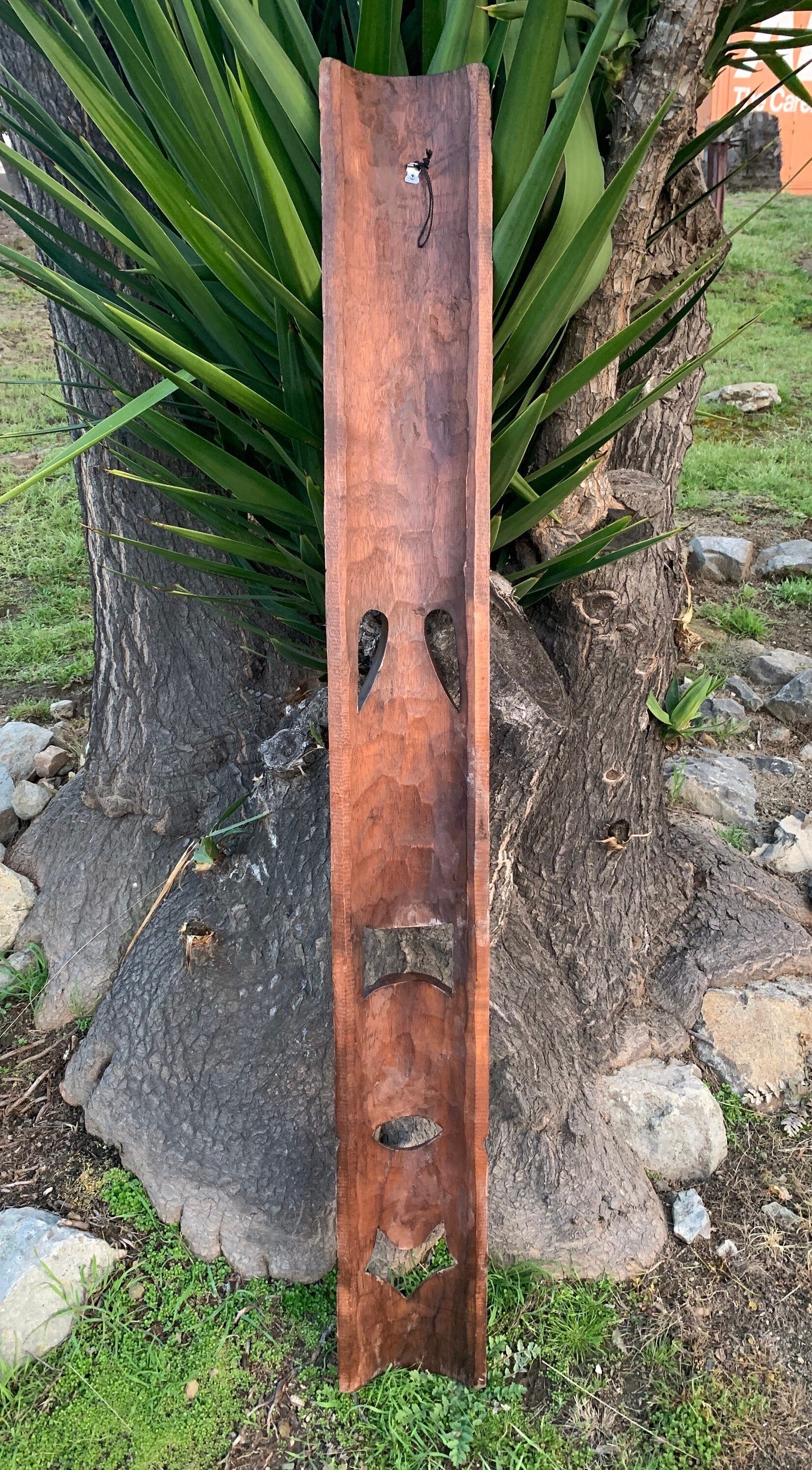 Tall carved wooden panel with geometric cutouts leaning against a tree in a garden.